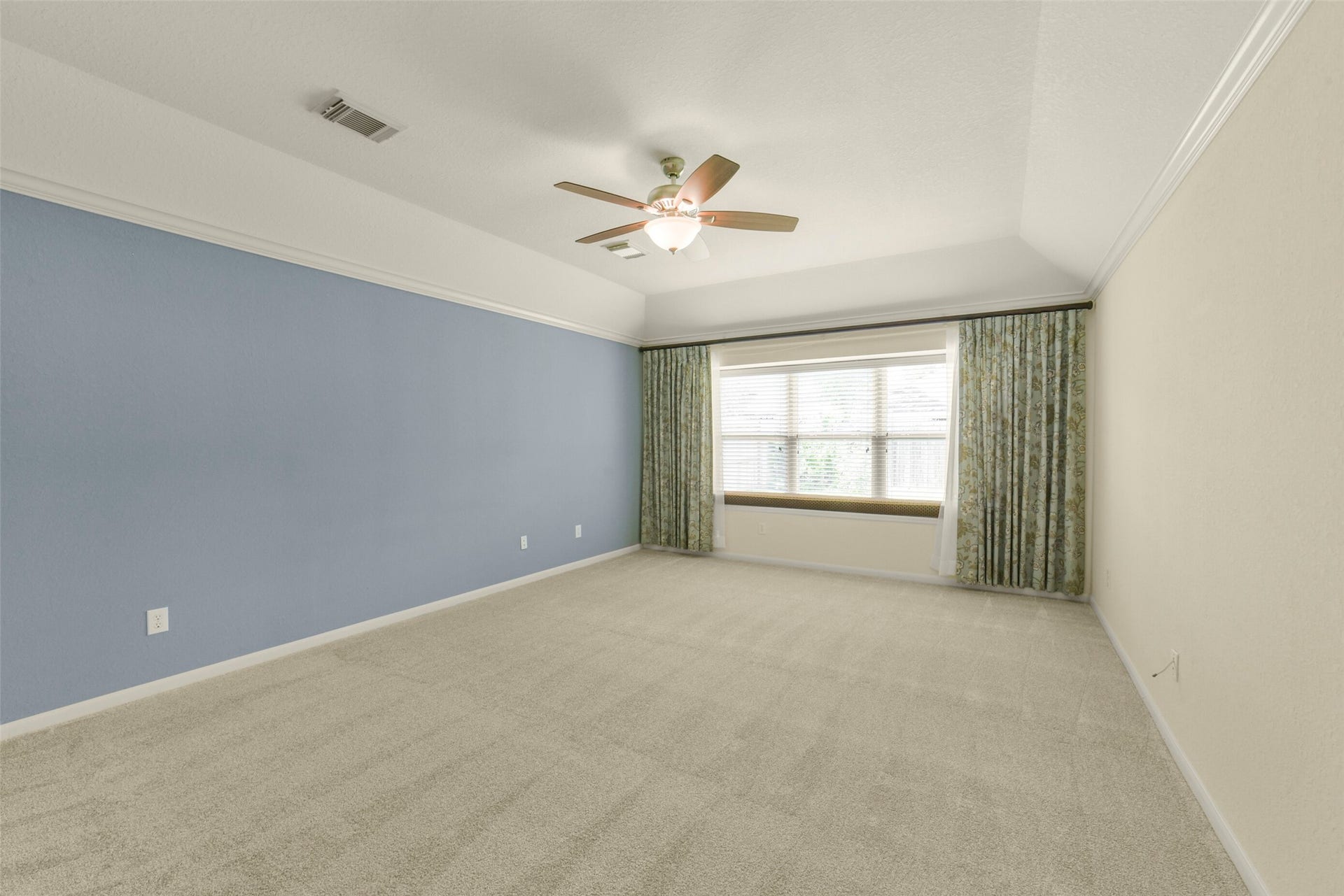 Spacious primary bedroom with soft carpet, a soothing blue accent wall, and a large window offering plenty of natural light, complemented by elegant ceiling fan and stylish drapery.