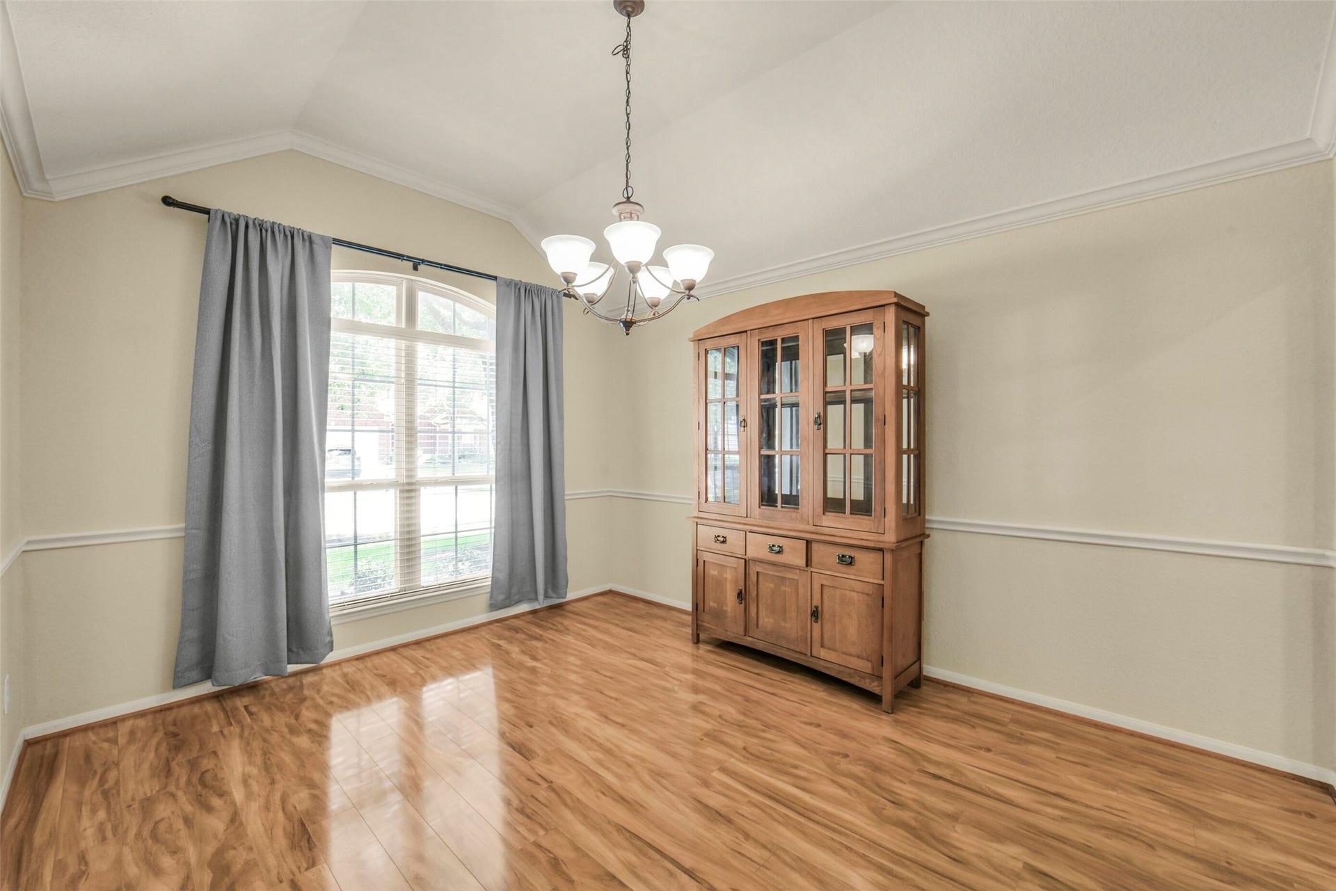 Bright and inviting dining room with elegant wood flooring, a charming chandelier, and a large window framed by tasteful curtains, perfect for gatherings.