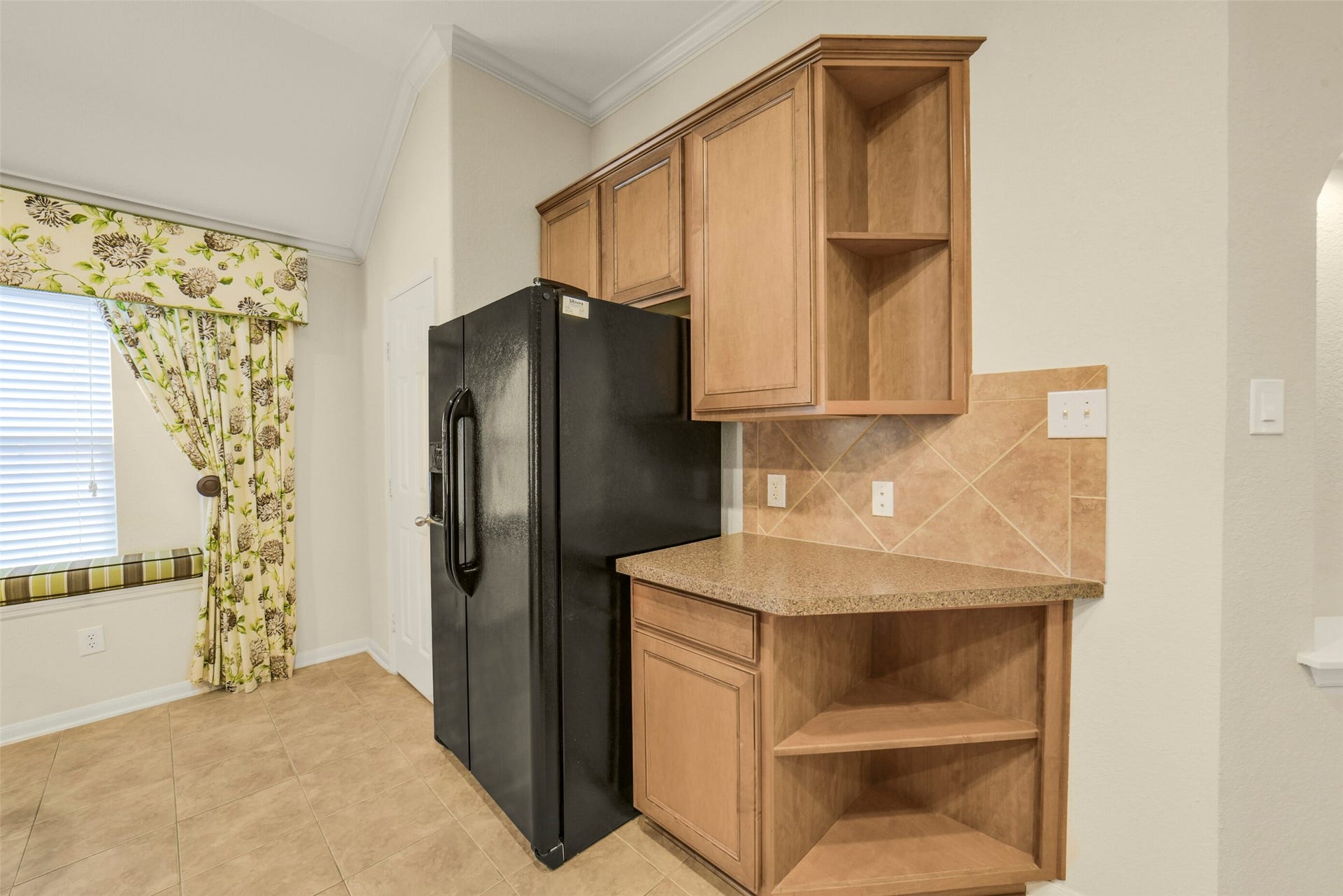 Charming kitchen corner with light wood cabinetry, sleek black refrigerator, and a cozy window seat adorned with floral drapes.