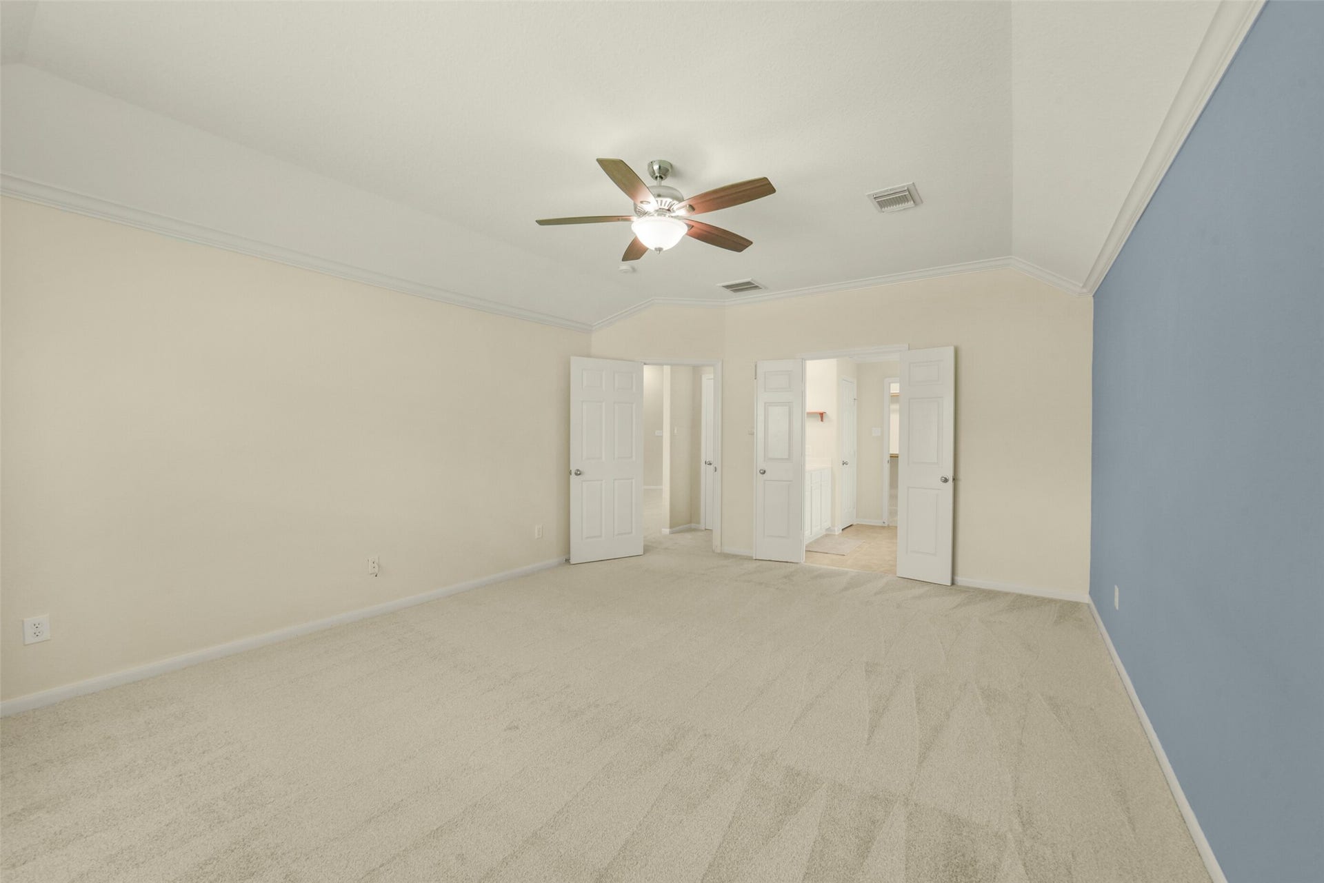 Spacious primary bedroom featuring plush carpeting, a soothing blue accent wall, and a ceiling fan, leading to an en-suite bathroom through double doors.