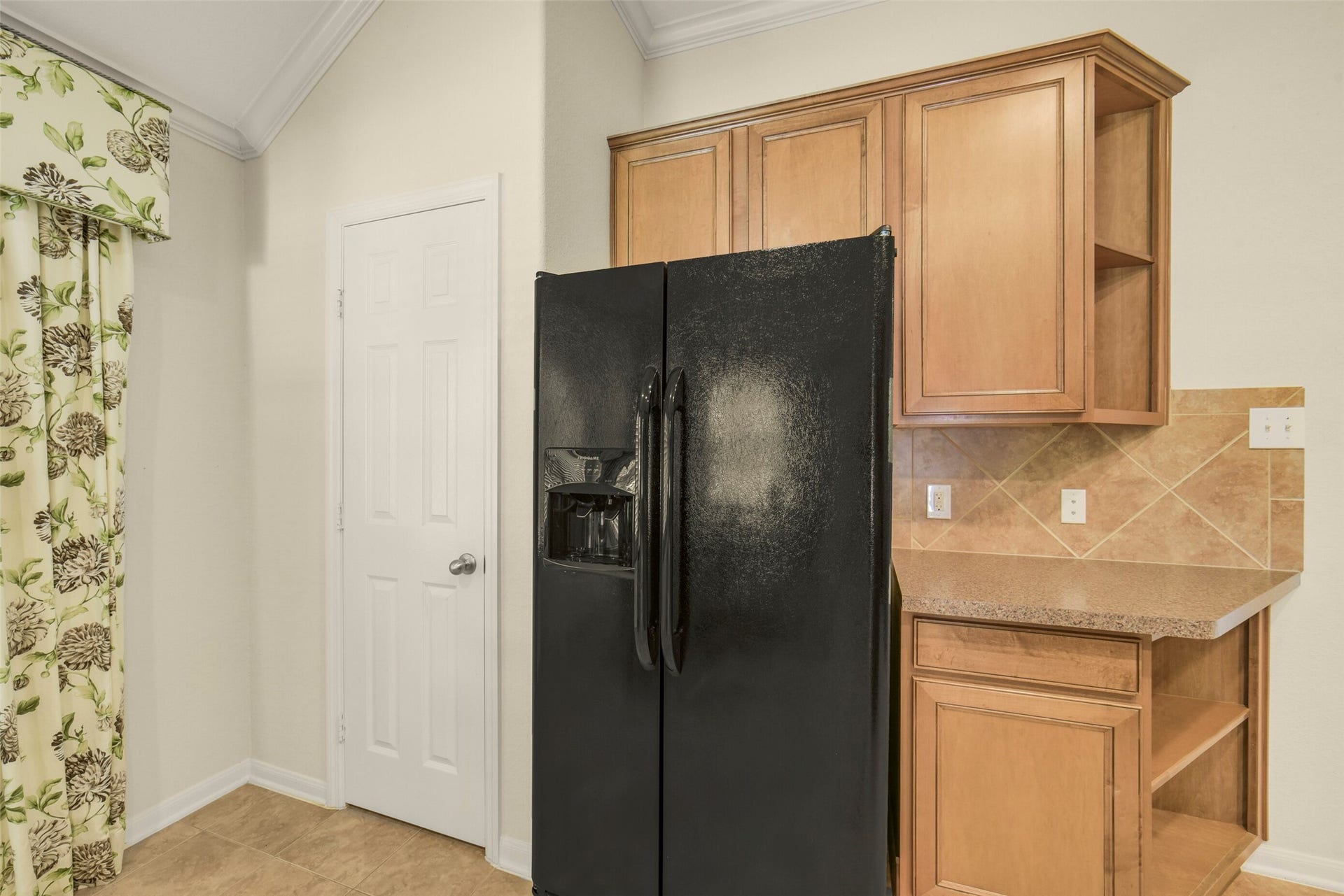 Spacious kitchen corner featuring a sleek black refrigerator, warm wood cabinetry, and elegant tile backsplash, complemented by stylish floral drapery.