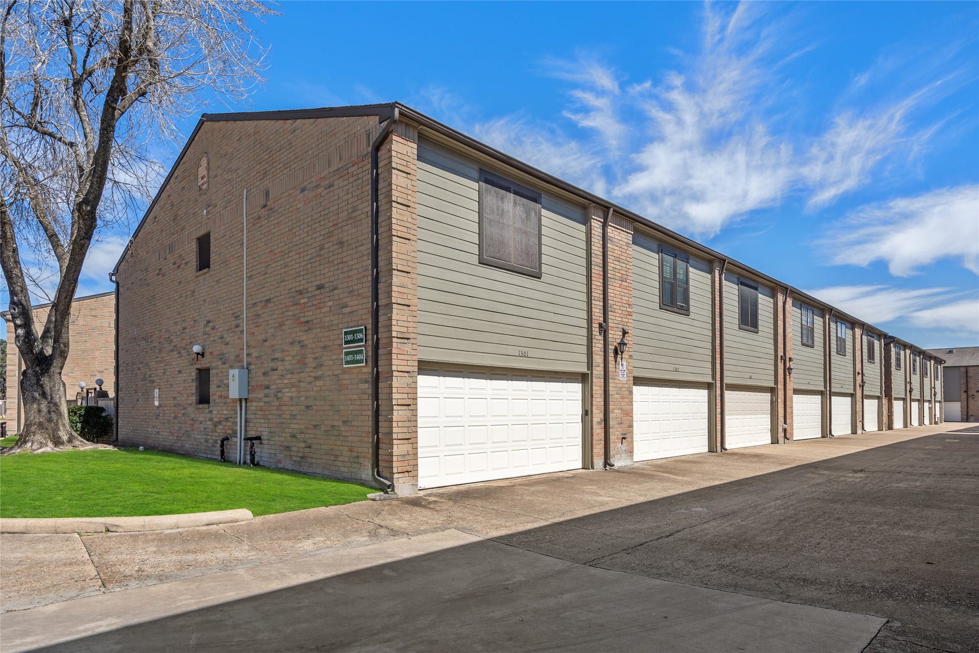 9850 Pagewood Lane, #1302, Houston, TX 77042 Rear view of the well-maintained townhome community, showcasing a row of two-car garages with clean white doors. The brick and siding exterior provides a classic and modern blend, while upper-level windows invite natural light inside. The paved driveway offers ample space for parking and easy access. A neatly landscaped green space with a mature tree adds a touch of nature to the area.