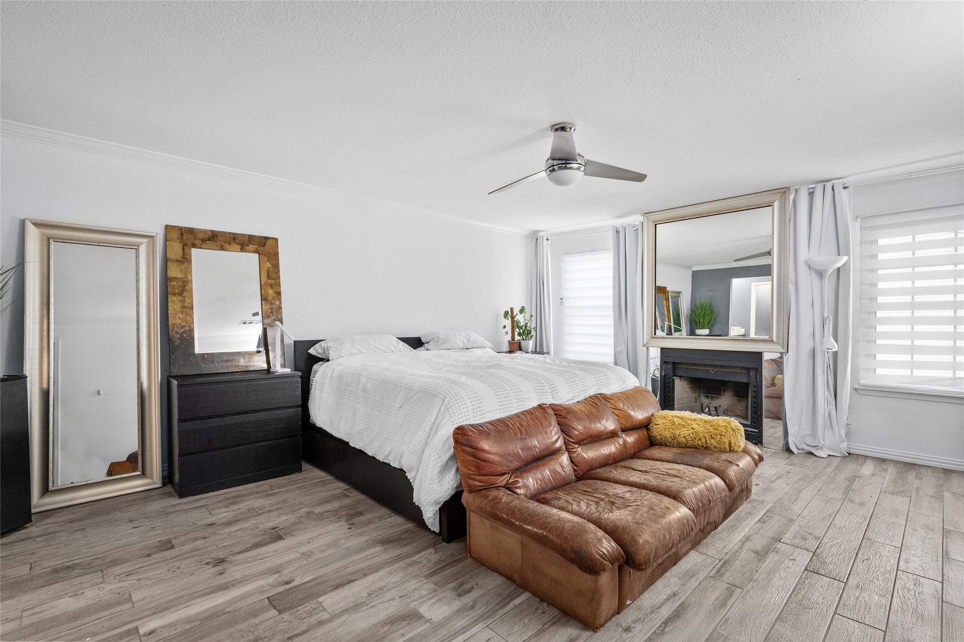 9850 Pagewood Lane, #1302, Houston, TX 77042 This expansive primary bedroom offers a serene retreat with its neutral tones, wood-look flooring, and abundant natural light. The cozy fireplace, framed by a statement mirror, adds warmth and character, while the modern ceiling fan ensures year-round comfort. Dual windows with sleek blinds provide adjustable privacy and brightness