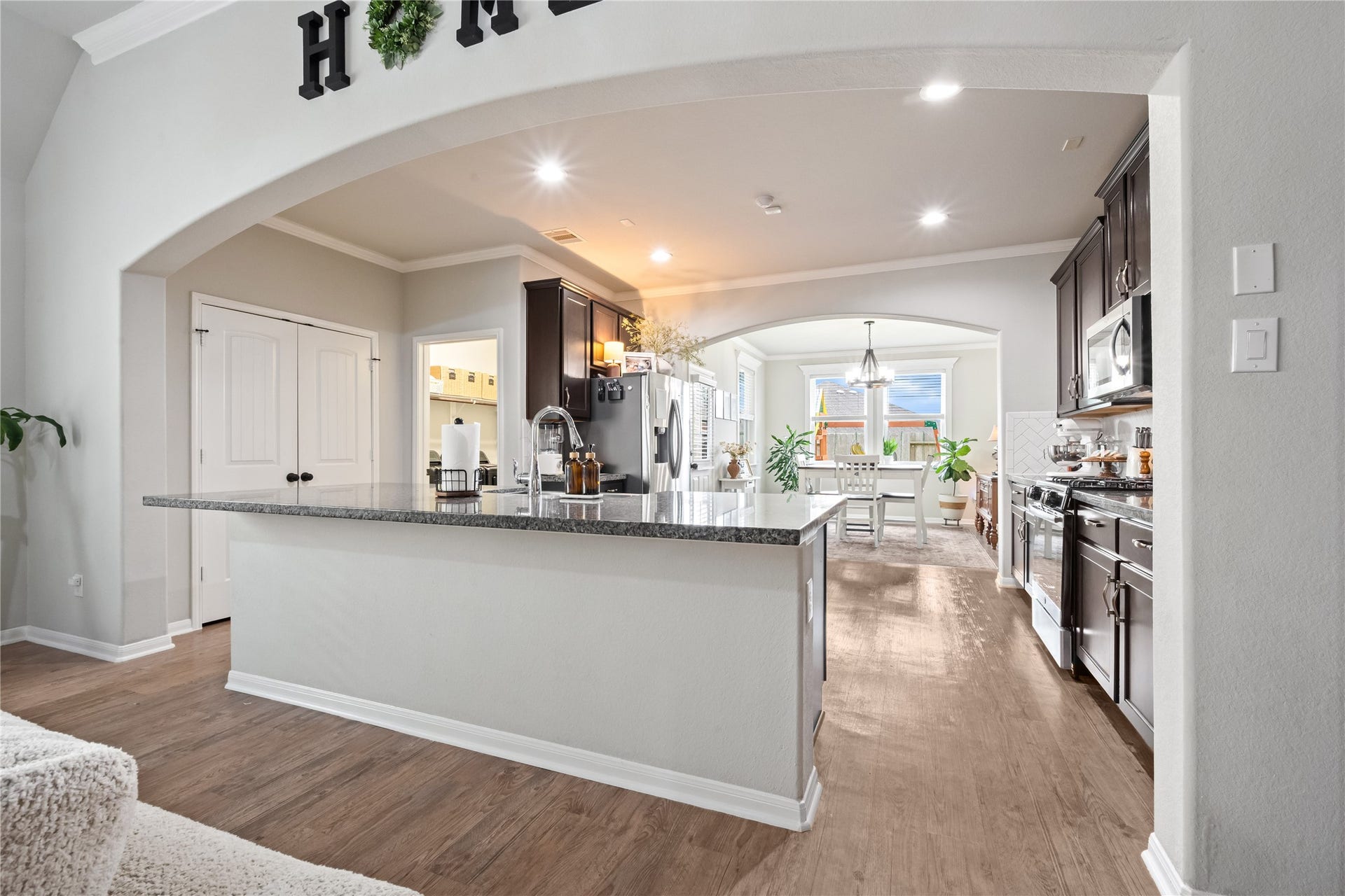 This photo showcases a modern kitchen with dark cabinetry, a large granite island, and stainless steel appliances. The open layout flows into a bright dining area, featuring wood flooring and elegant archways. Perfect for entertaining and family gatherings.