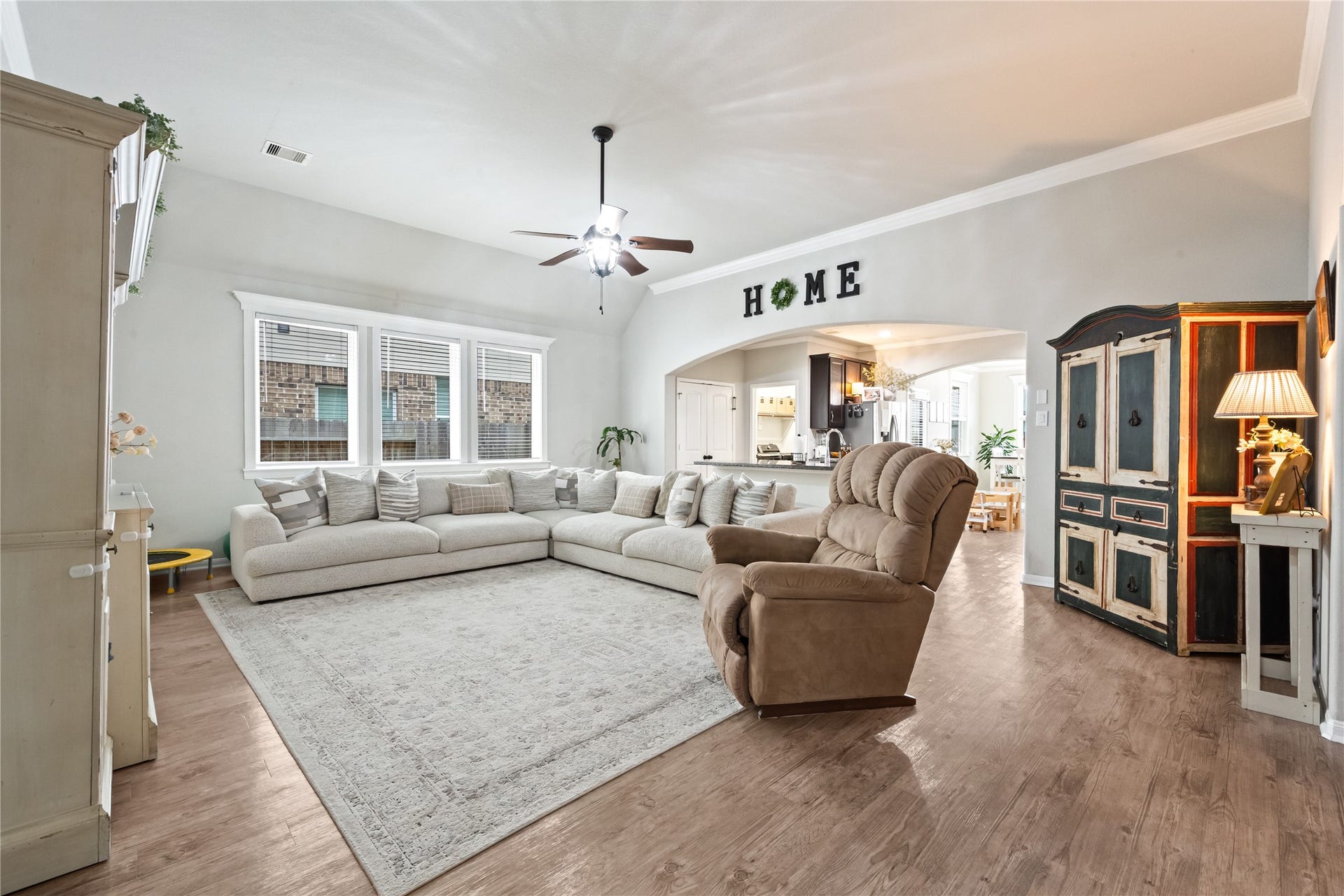 Spacious living room with high ceilings, a large sectional sofa, and a cozy recliner. It features warm wood flooring, abundant natural light from multiple windows, and an open layout leading into the kitchen. Perfect for family gatherings.