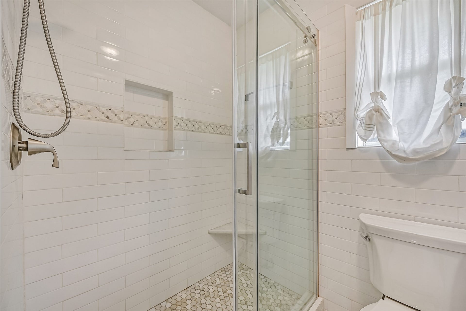304 Chester Drive, Friendswood, TX 77546 Master Bathroom First Residence:
Walk-in glass-enclosed shower featuring classic white subway tile, a decorative mosaic accent band, built-in niche and bench, hex tile flooring, and a window providing natural light for a bright, spa-inspired feel.