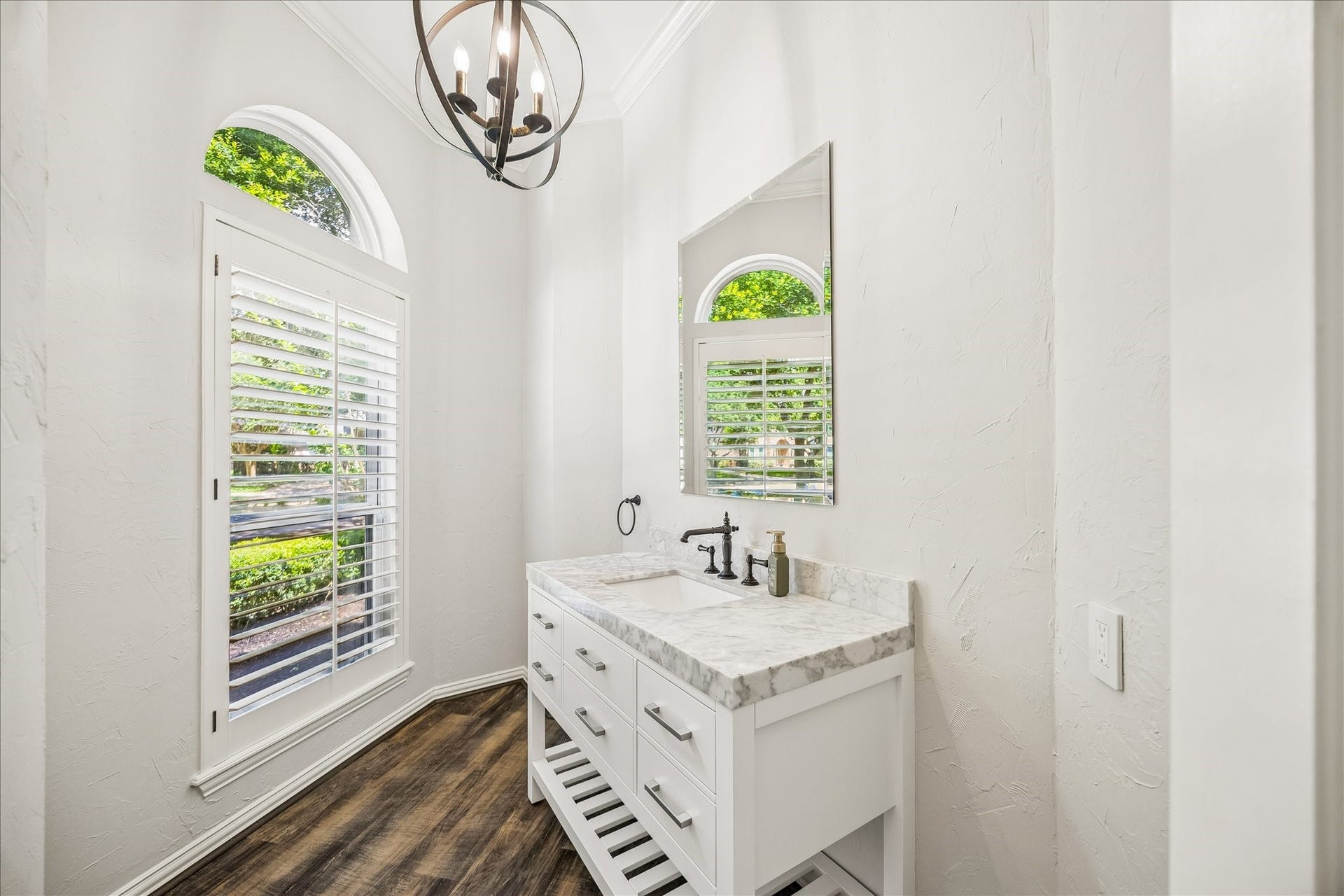 This light-filled half bath showcases a marble countertop vanity with refined designer fixtures, complemented by an expansive window dressed with plantation shutters that flood the space with natural light. Sophisticated wood-style flooring adds warmth and elegance, creating a polished, upscale finish.