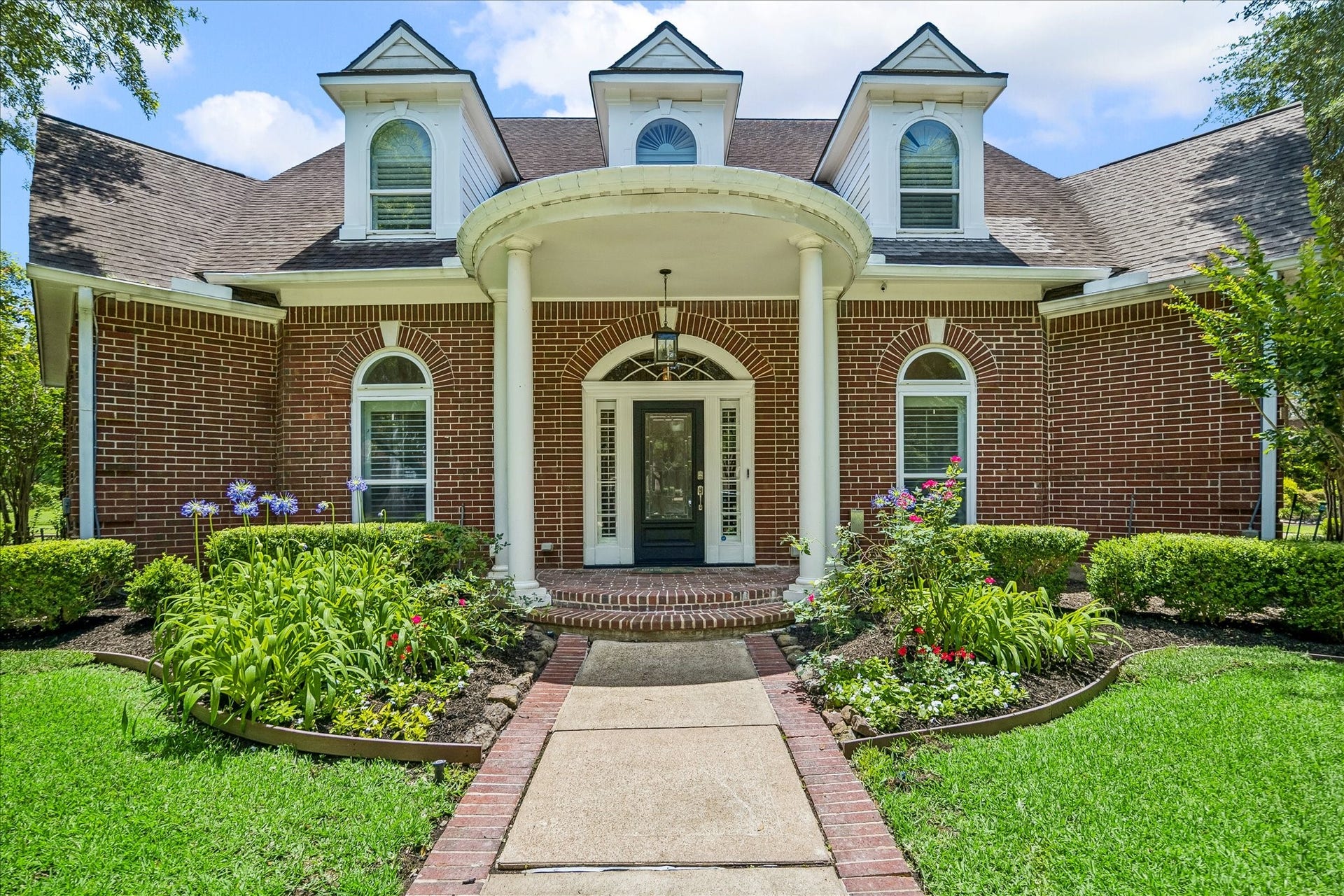 This charming two-story brick home with dormer windows and a welcoming front porch, is set on a spacious, lush green lawn framed by mature trees. The property features a curved walkway leading to the entrance, creating a picturesque and inviting curb appeal.