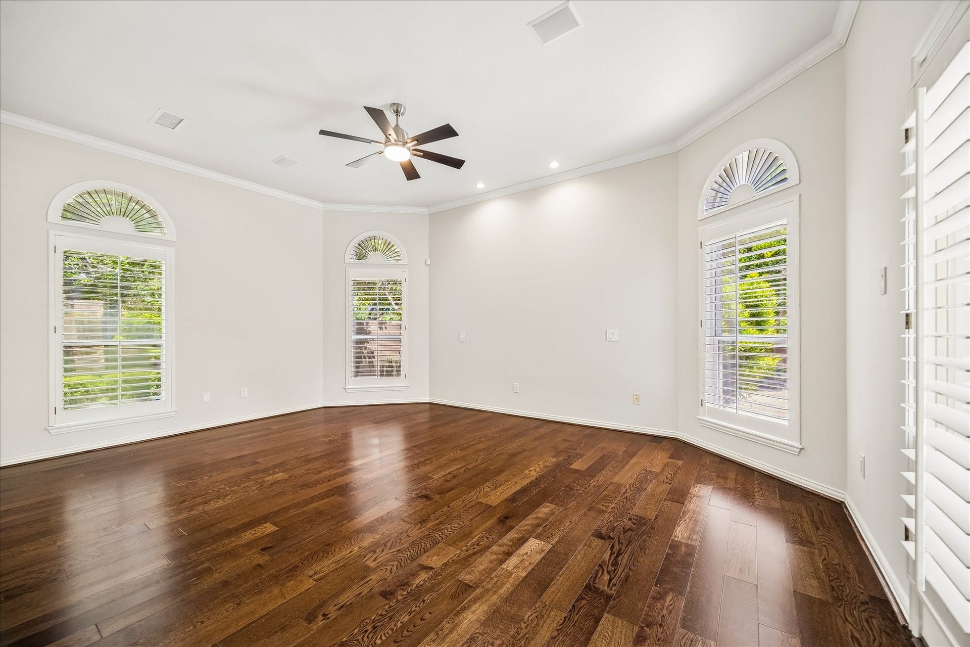 This bright and spacious primary suite showcases rich hardwood floors, large arched windows with plantation shutters, and a ceiling fan. Thoughtfully designed for comfort and style, the room offers a warm, inviting atmosphere with timeless elegance.