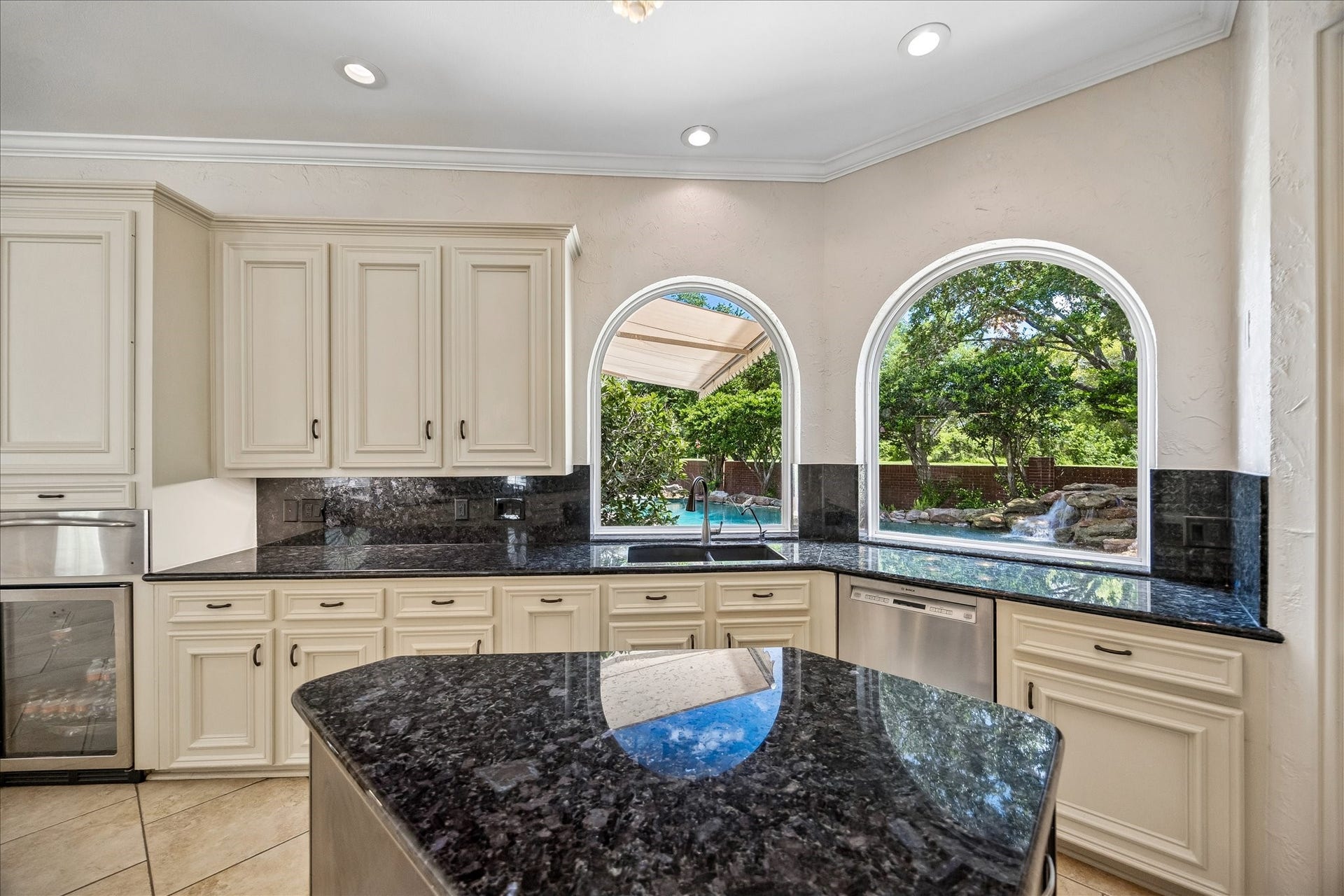 This kitchen features elegant cream colored cabinetry with granite countertops. Two large arched windows offer a view of a lush backyard with a pool and waterfall, providing a serene backdrop.