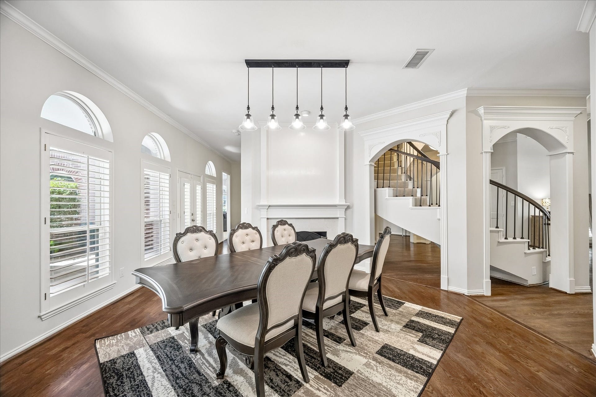 Elegant dining room featuring grand arched windows, rich hardwood flooring, and a striking modern chandelier. The space blends timeless architectural details with contemporary sophistication and is anchored by a stunning double-sided fireplace.
