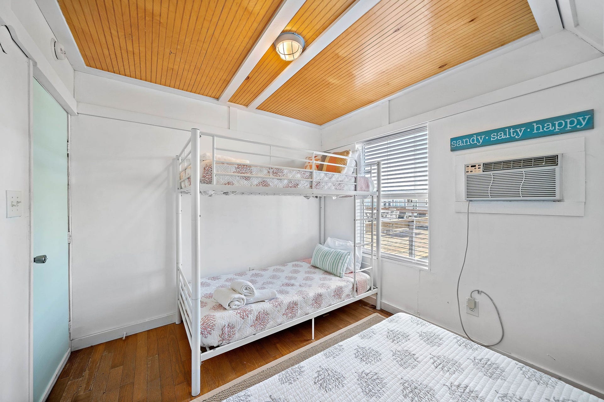 16610 Nassau Way, Galveston, TX 77554 Bright and inviting bedroom featuring a stylish bunk bed, wood-paneled ceiling, and a large window for natural light. A wall-mounted air conditioner ensures comfort, while a cheerful decor sign adds a playful touch.