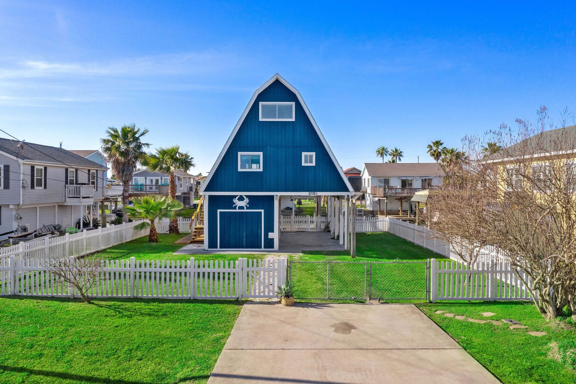16610 Nassau Way, Galveston, TX 77554 Charming A-frame blue house with a white picket fence, spacious driveway, and lush green lawn, offering a coastal feel with palm trees and a welcoming front porch.