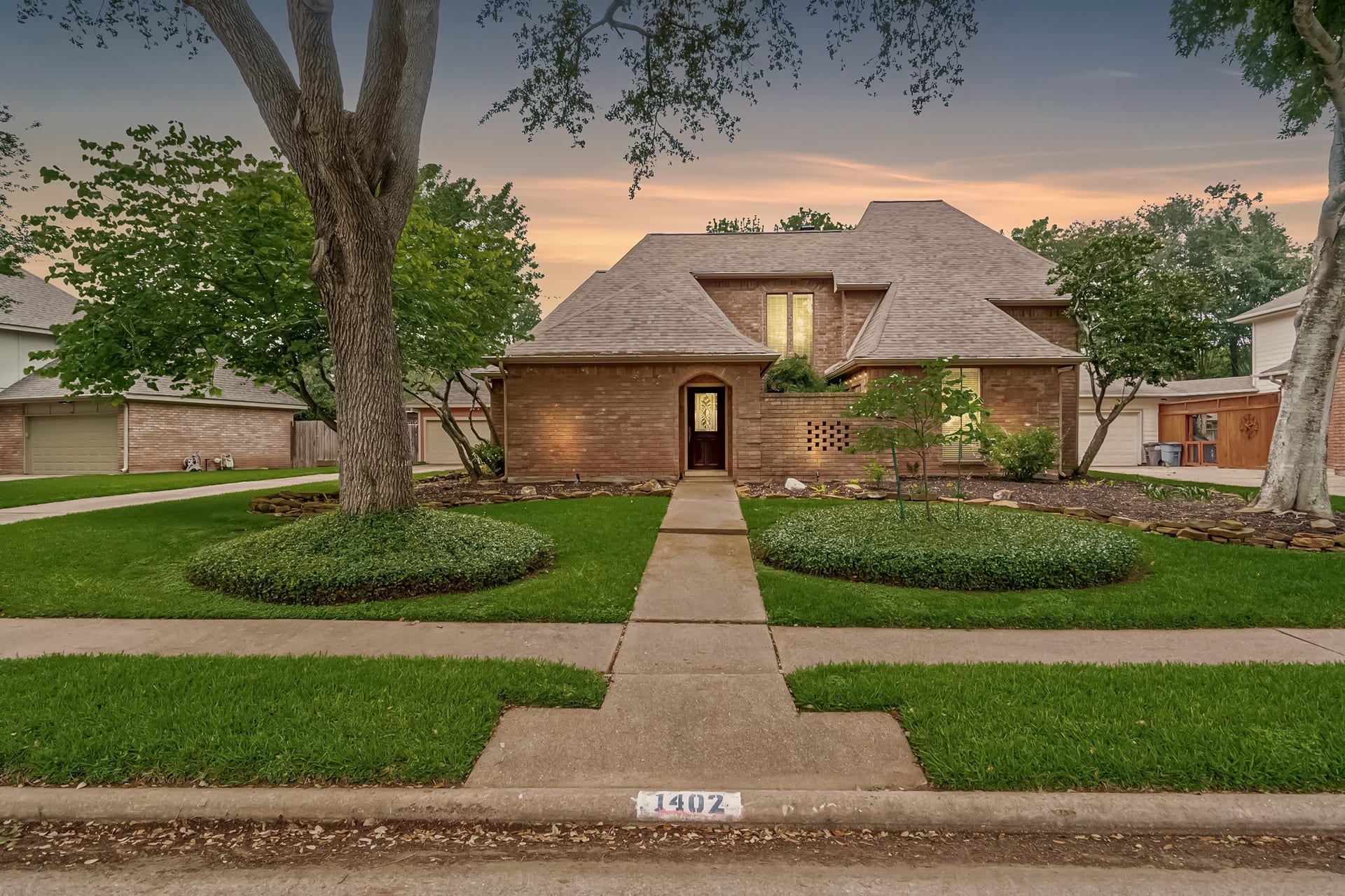 A welcoming first impression of 1402 Bittersweet Drive, showcasing the traditional brick exterior and manicured curb appeal under a soft sunset sky. The symmetrical front walkway and mature landscaping highlight the pride of ownership in this Pecan Grove Plantation home.