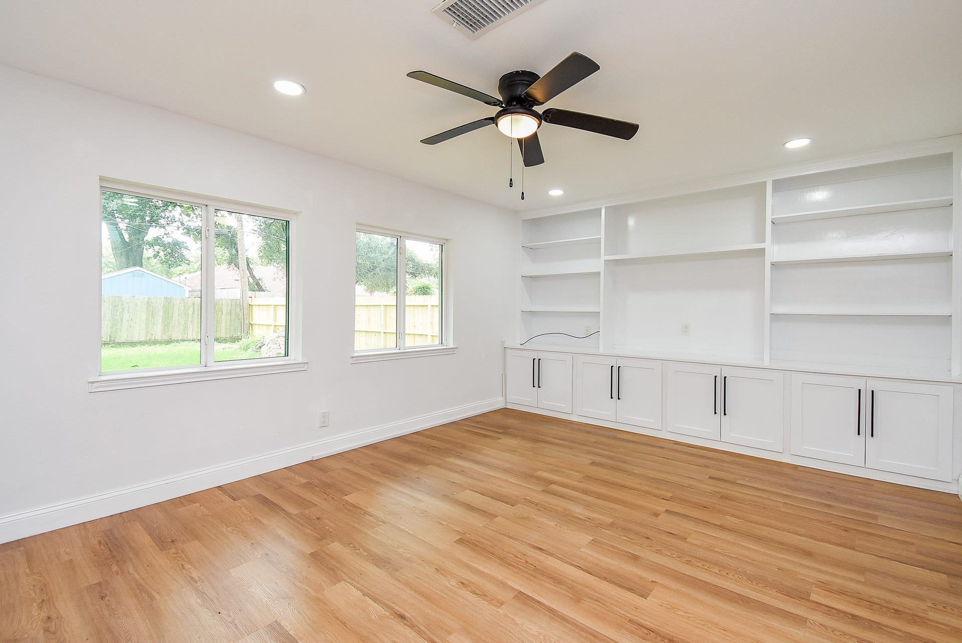 Bright room with built-in shelving, wood floors, large backyard-facing windows, and a ceiling fan.