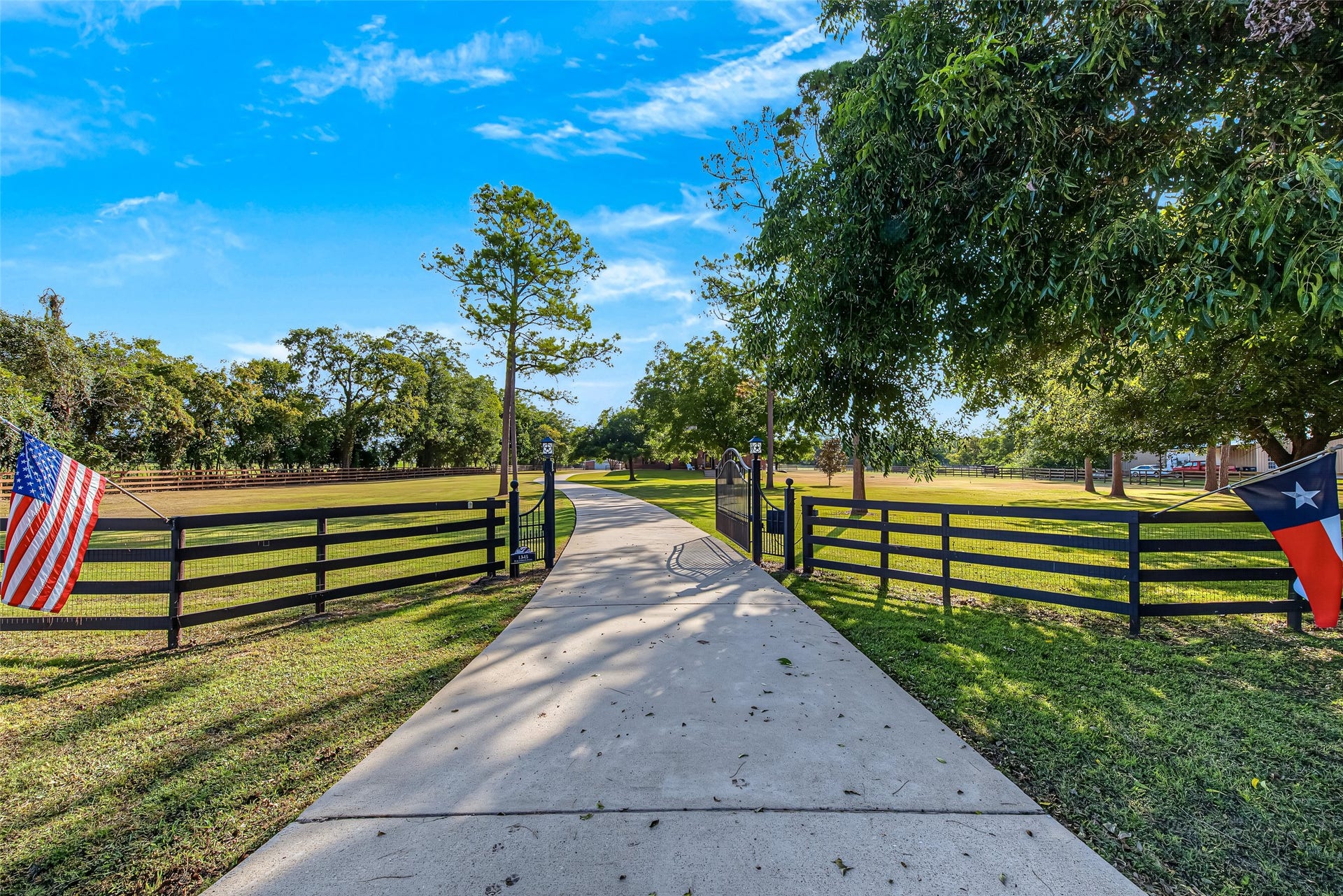 1345 Peach Ridge Road, Brookshire, TX 77423 Creating an immediate sense of arrival, the estate entry is framed by a private wrought-iron automatic security gate anchored by dark wood-and-wire post fencing along the front perimeter. A clean concrete driveway winds through immaculate green grounds shaded by towering mature trees, proudly flanked by American and Texas flags that welcome you to the property.