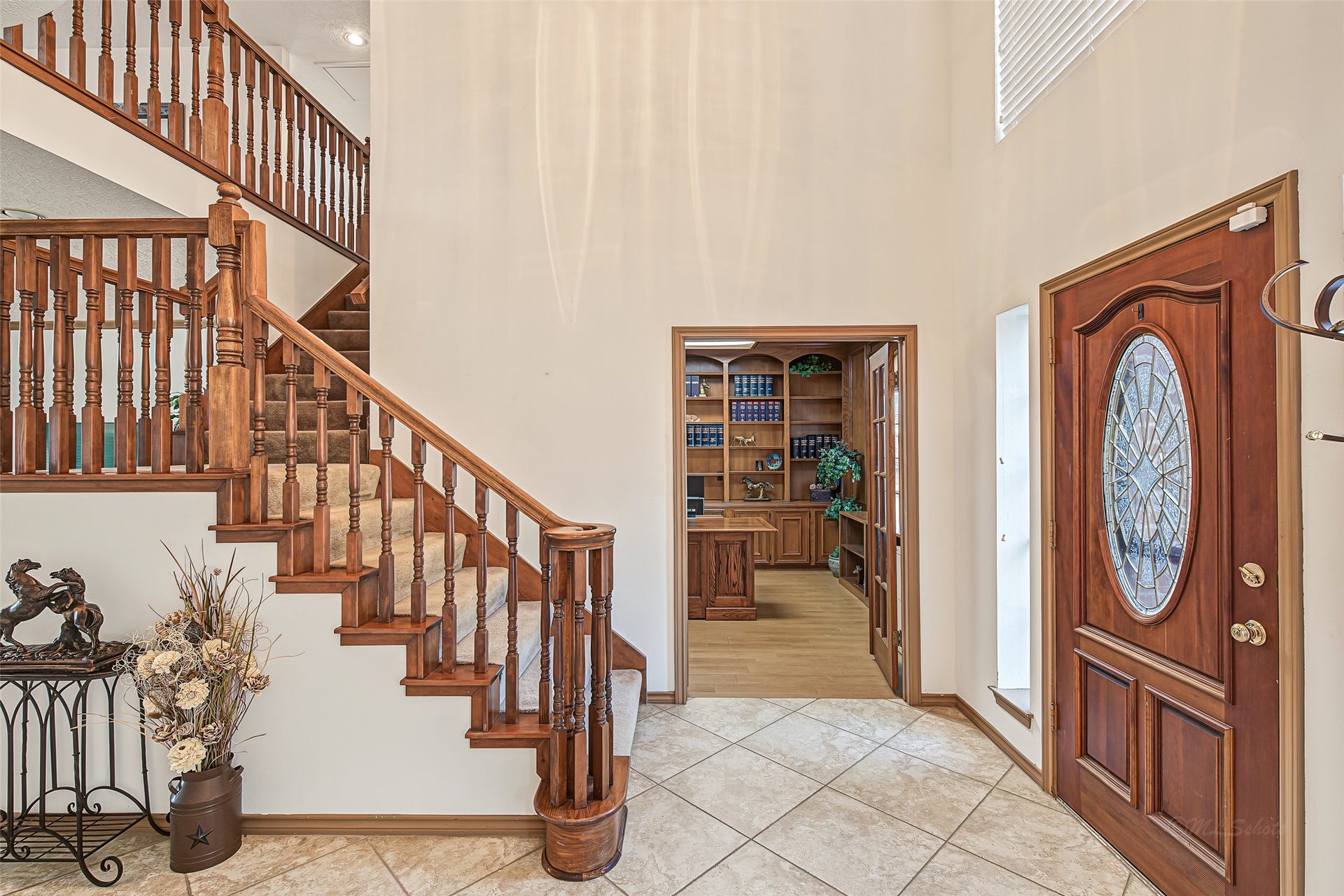 1345 Peach Ridge Road, Brookshire, TX 77423 This grand, two-story foyer makes a striking first impression with its soaring high ceilings, anchored by a solid wood front door featuring an ornate oval leaded-glass insert. The entryway showcases a beautiful open staircase crafted with detailed wood balusters and a graceful carved newel post. Diagonal-set ceramic tile flooring guides guests effortlessly through the space, framing an inviting view straight into the private study.