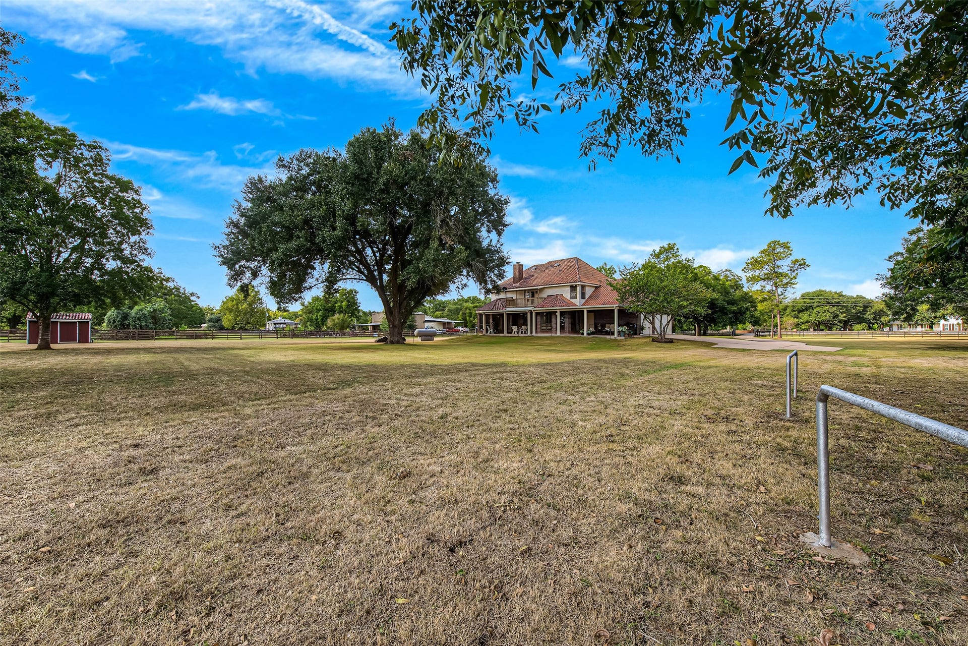 1345 Peach Ridge Road, Brookshire, TX 77423 Showcasing true country estate living, this expansive property offers picturesque acreage surrounded by mature hardwood shade trees and open green pastures. The two-story residence is framed by a sweeping wraparound porch and second-story balcony, setting a classic, peaceful backdrop complete with perimeter pipe fencing and a charming red storage shed or barn structure in the distance.