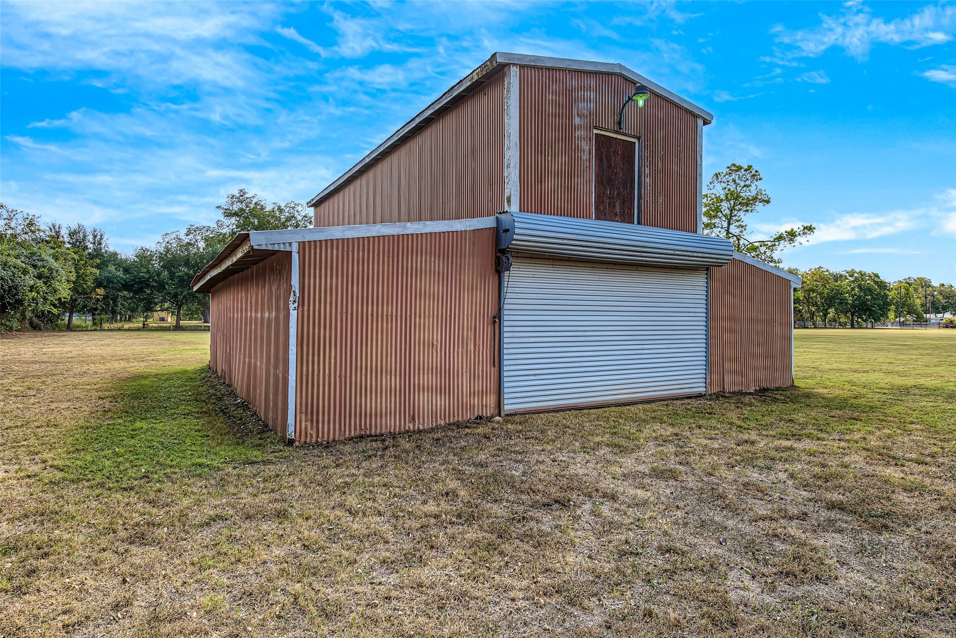 1345 Peach Ridge Road, Brookshire, TX 77423 A standout asset for country living, this substantial freestanding metal barn/workshop features durable corrugated metal siding, a wide metal roll-up door for easy vehicle or equipment entry, and a upper loft level complete with top-door access and exterior gooseneck lighting. Enclosed by lean-to side wings for additional covered workspace or storage, this versatile structure is ideal for housing tractor equipment, workshop tools, boats, RV storage, or agricultural supplies.