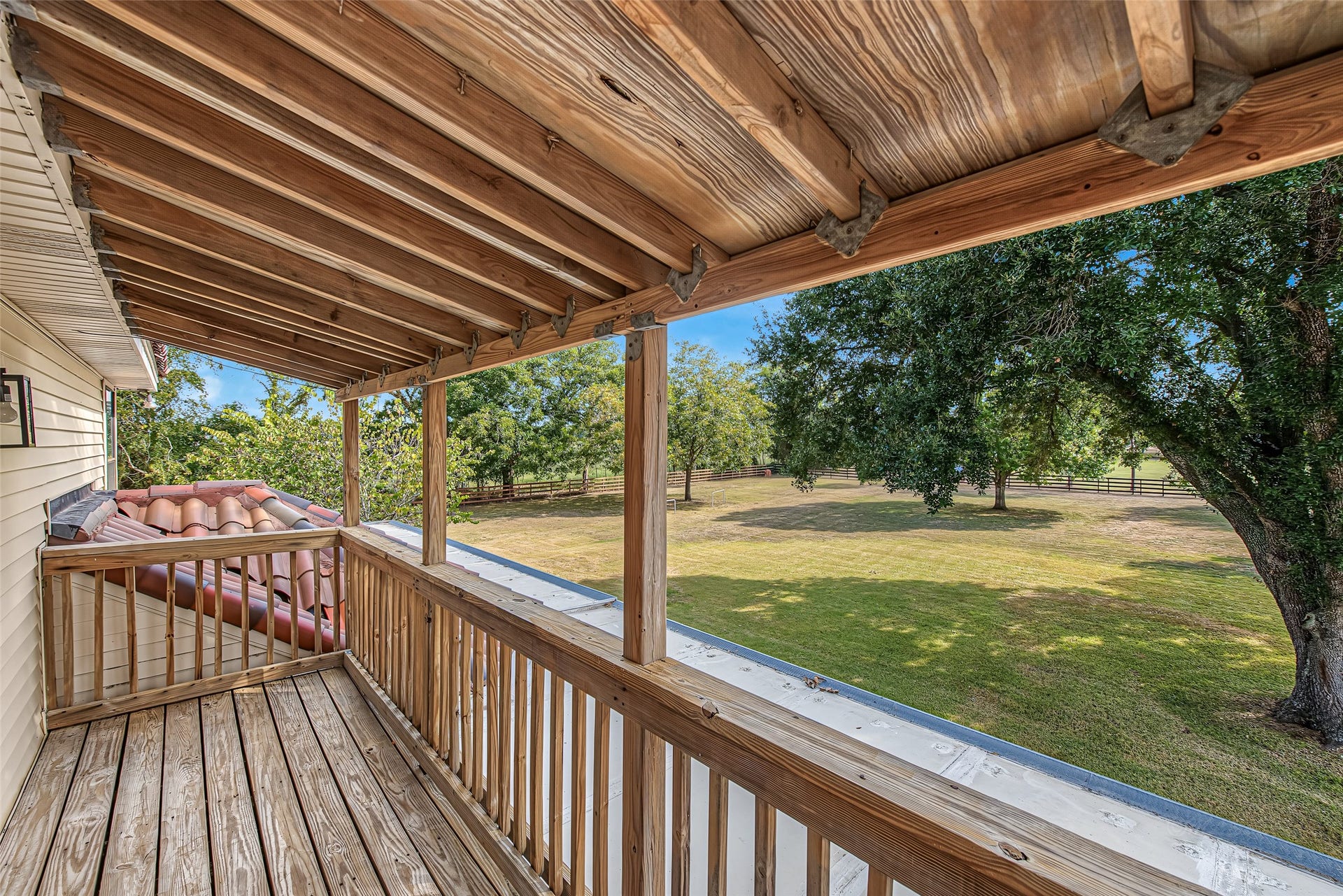 1345 Peach Ridge Road, Brookshire, TX 77423 Offering an elevated vantage point over the property, this covered second-story wood balcony features exposed timber ceiling joists, wood plank decking, and a sturdy perimeter railing. Directly accessible from the upper living quarters, this private outdoor retreat is shaded for year-round comfort while capturing panoramic views of the lush green acreage, mature shade trees, and surrounding fenced pastures.