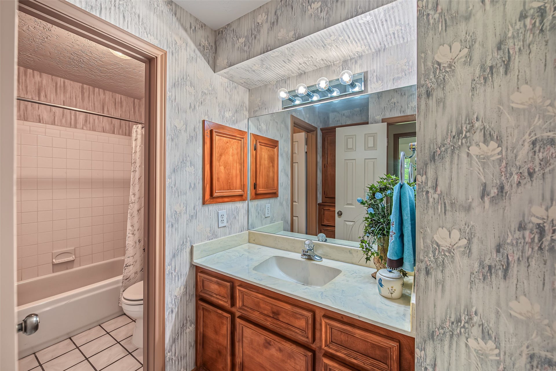 1345 Peach Ridge Road, Brookshire, TX 77423 This well-designed secondary bathroom features a functional split-vanity layout that separates the primary grooming space from the tub and toilet area. The vanity zone offers a solid wood vanity with raised-panel cabinetry, a light marble-look countertop with an undermount sink, a large wall-to-wall mirror, and overhead vanity strip lighting.