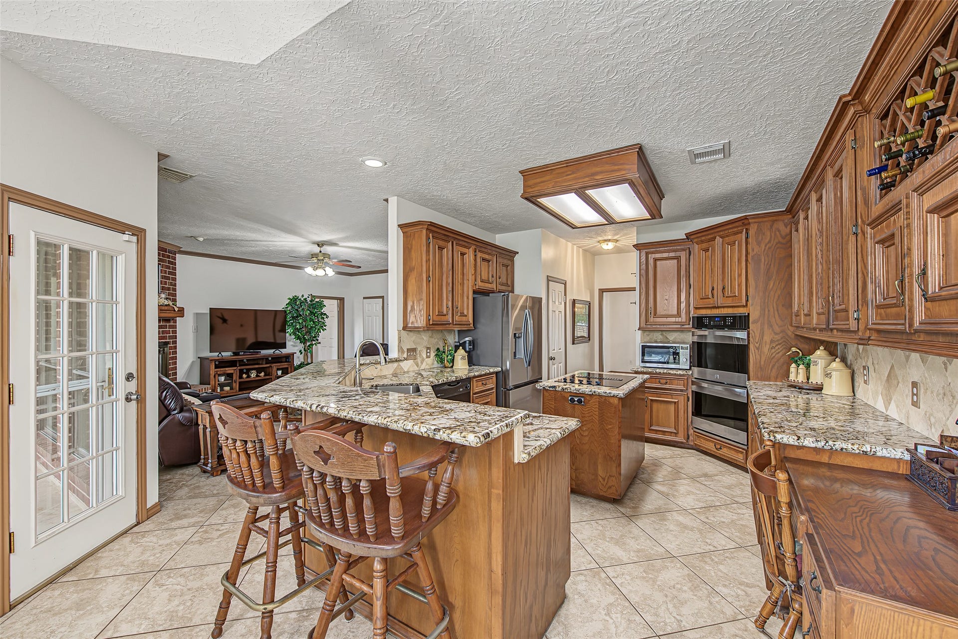 1345 Peach Ridge Road, Brookshire, TX 77423 An elevated breakfast bar with barstool seating naturally bridges the space into the adjacent living room, while custom details like an integrated wine rack, a built-in desk, and a multi-pane glass door leading outside enhance both form and function.