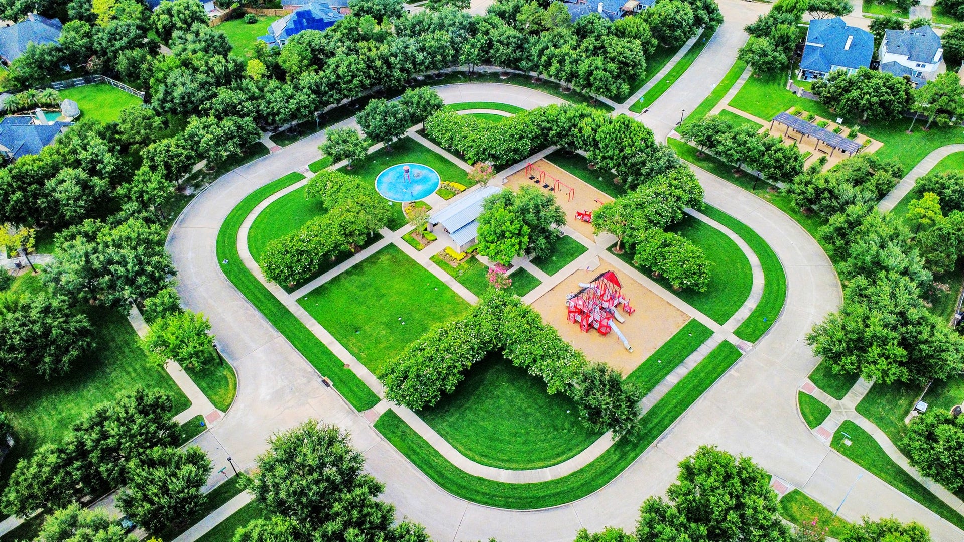 21123 Dover Park Lane, Katy, TX 77450 This aerial photo showcases a vibrant neighborhood park surrounded by a circular street. The park features a playground, open green spaces, and a splash pad, providing a family-friendly environment. Lush trees line the streets, and well-maintained homes are visible around the perimeter, indicating a tranquil and inviting residential area.