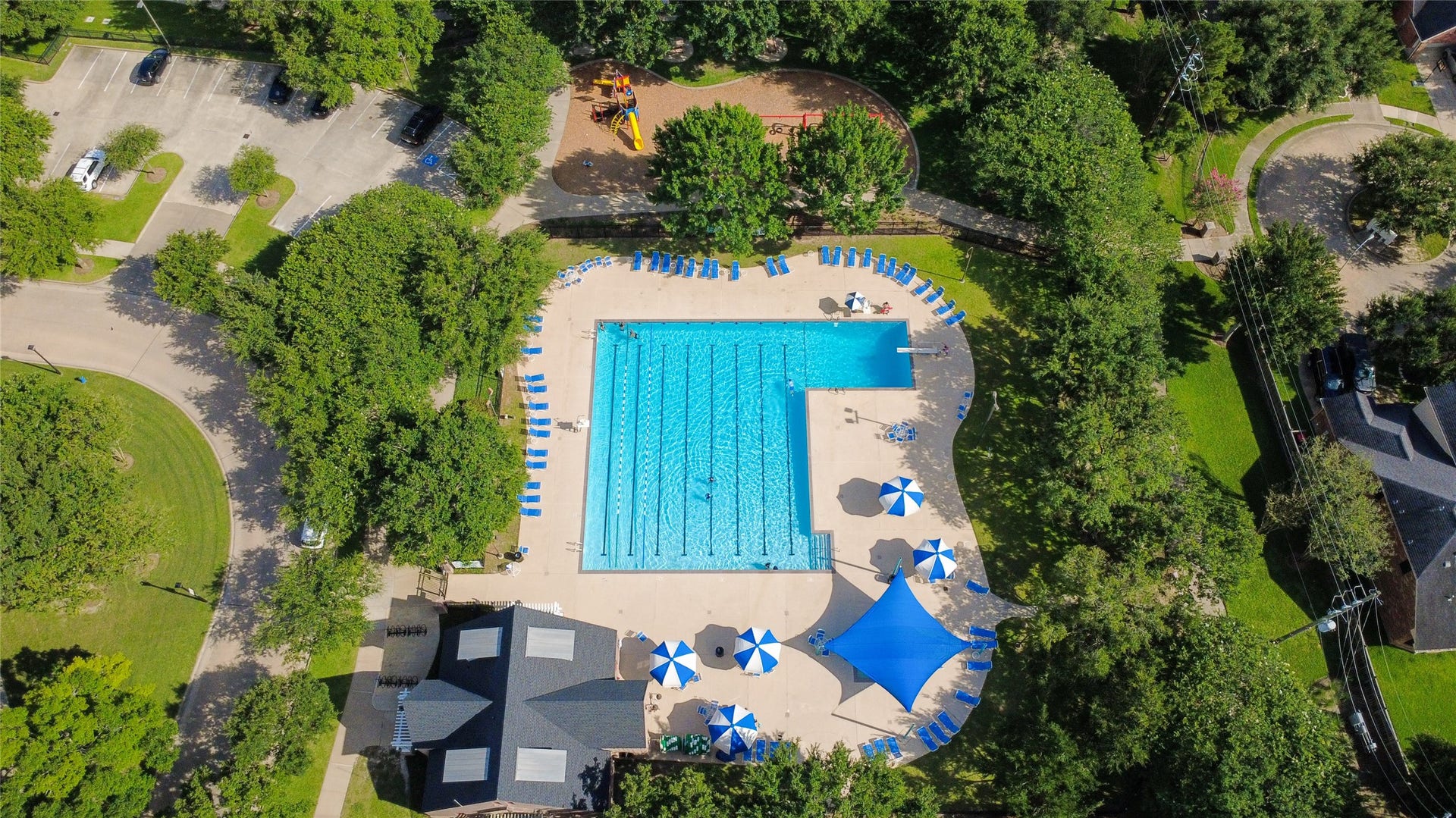 21123 Dover Park Lane, Katy, TX 77450 This aerial view showcases a well-maintained community pool area surrounded by lush greenery. It features a large swimming pool, ample lounge chairs, umbrellas, a shaded pavilion, and a nearby playground, offering a perfect recreational spot for residents.