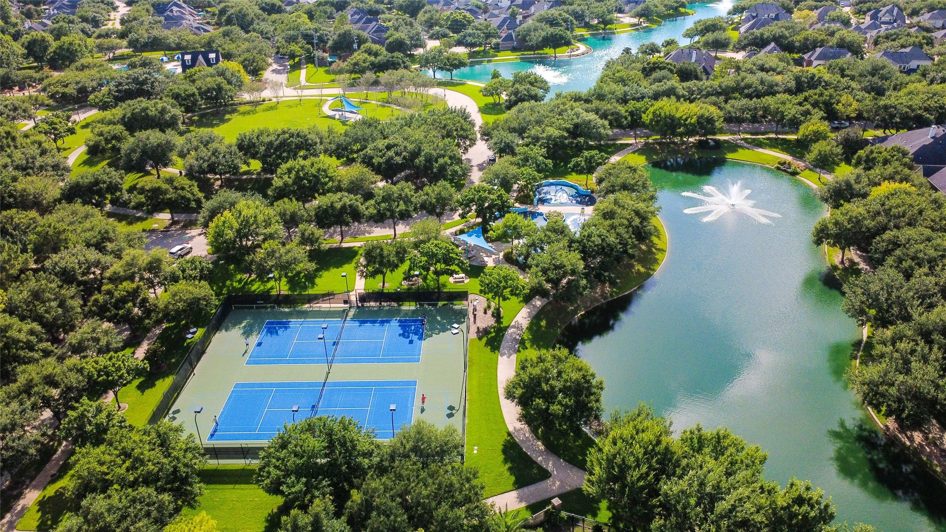21123 Dover Park Lane, Katy, TX 77450 This aerial photo showcases a picturesque residential community featuring lush greenery, two tennis courts, winding walking paths, a serene pond with a fountain, and a swimming pool. Ideal for active lifestyles and relaxation.