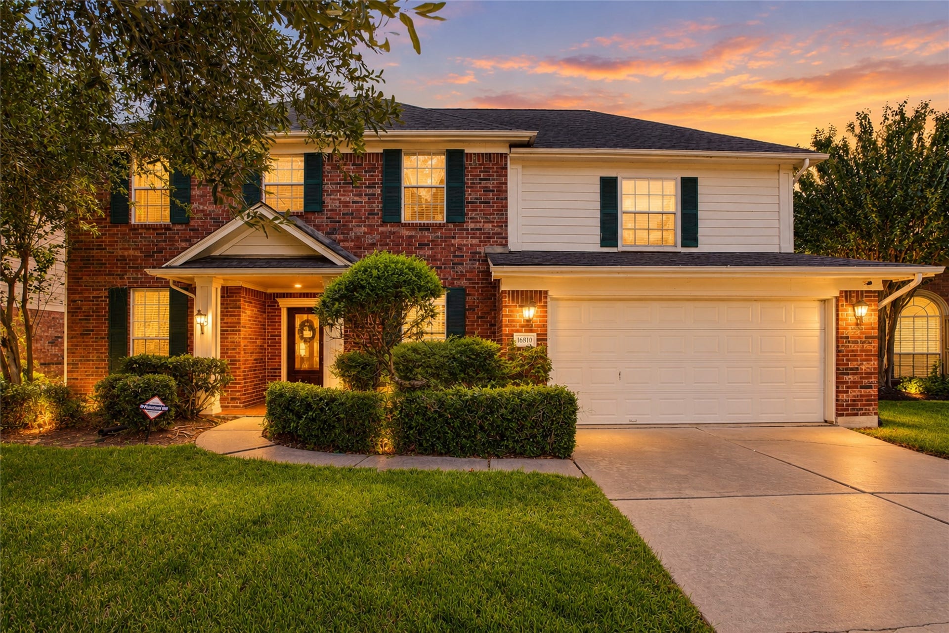 Two-story traditional with brick and siding exterior, dark shutters, manicured shrubs, mature trees, and an attached two-car garage. Digitally enhanced twilight image; landscape lighting shown is not present on the property.