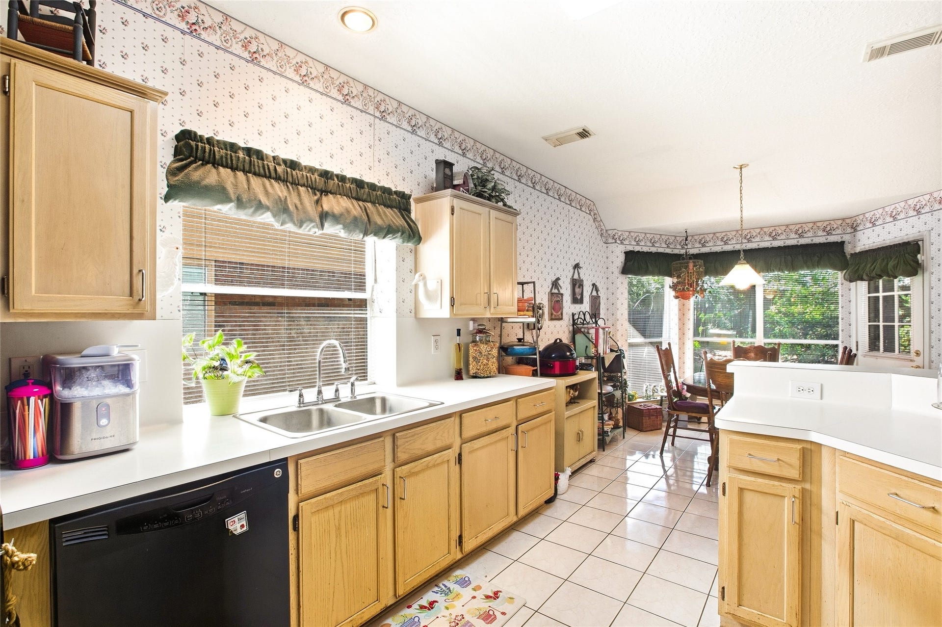 Kitchen sink wall with a double stainless sink under the window, light wood cabinetry, white countertops, and a black dishwasher, with views into the breakfast area.