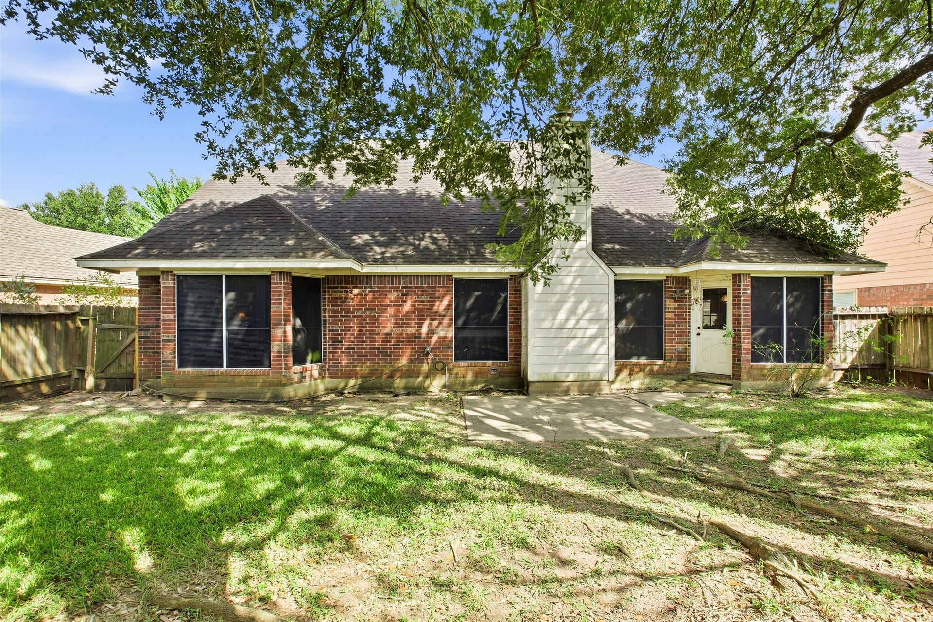 Rear exterior with brick construction, concrete patio, and wood privacy fence under a mature tree canopy.