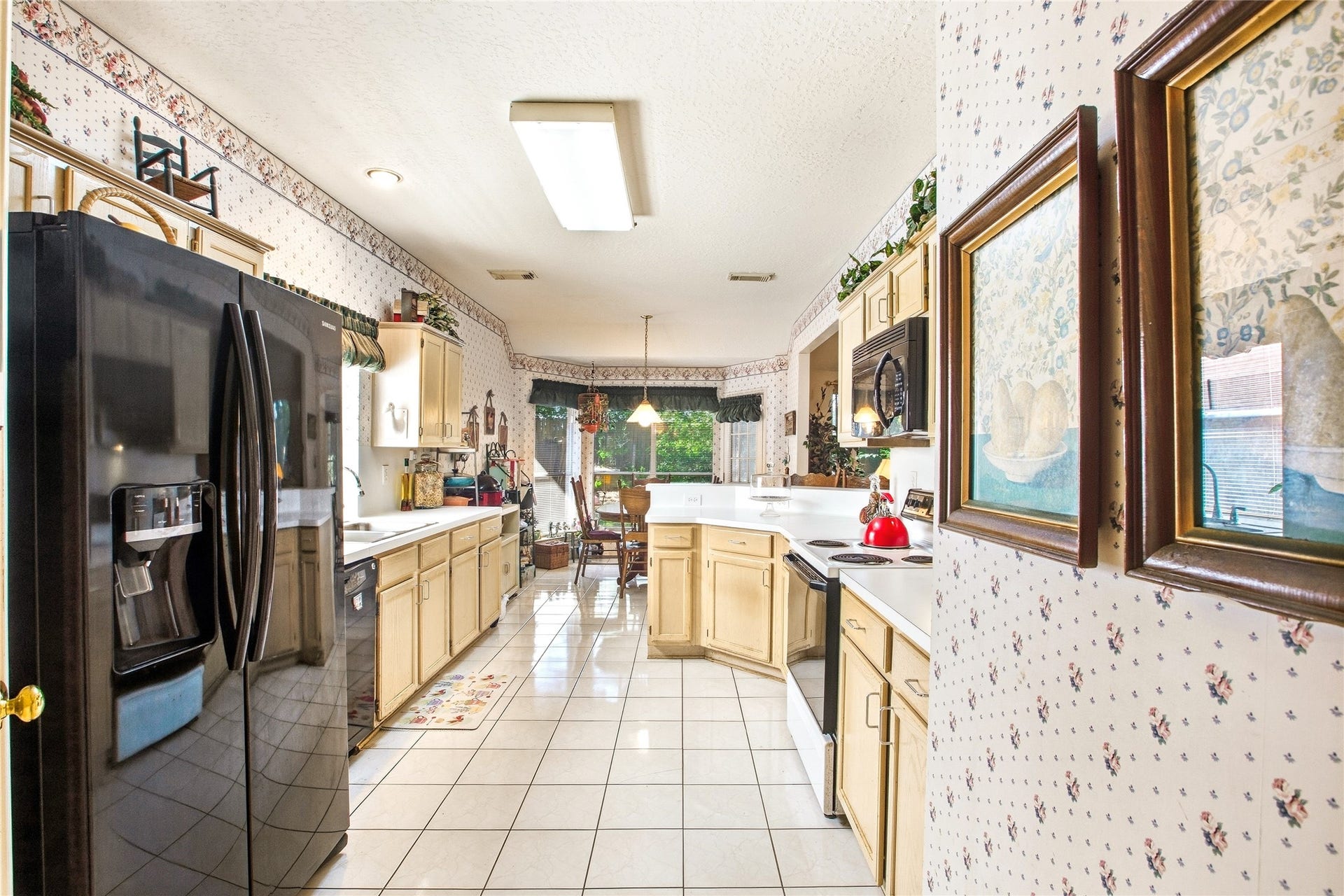 Kitchen with light wood cabinetry, white countertops, ceramic tile flooring, and a black side-by-side refrigerator, opening to the breakfast area with a bay window.