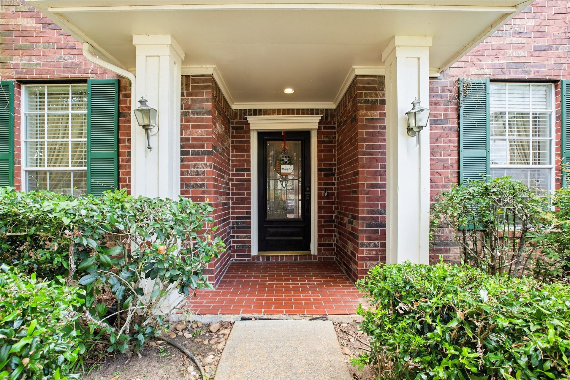 Covered front porch with white columns, brick-paver flooring, coach lights, and a front door with decorative glass.