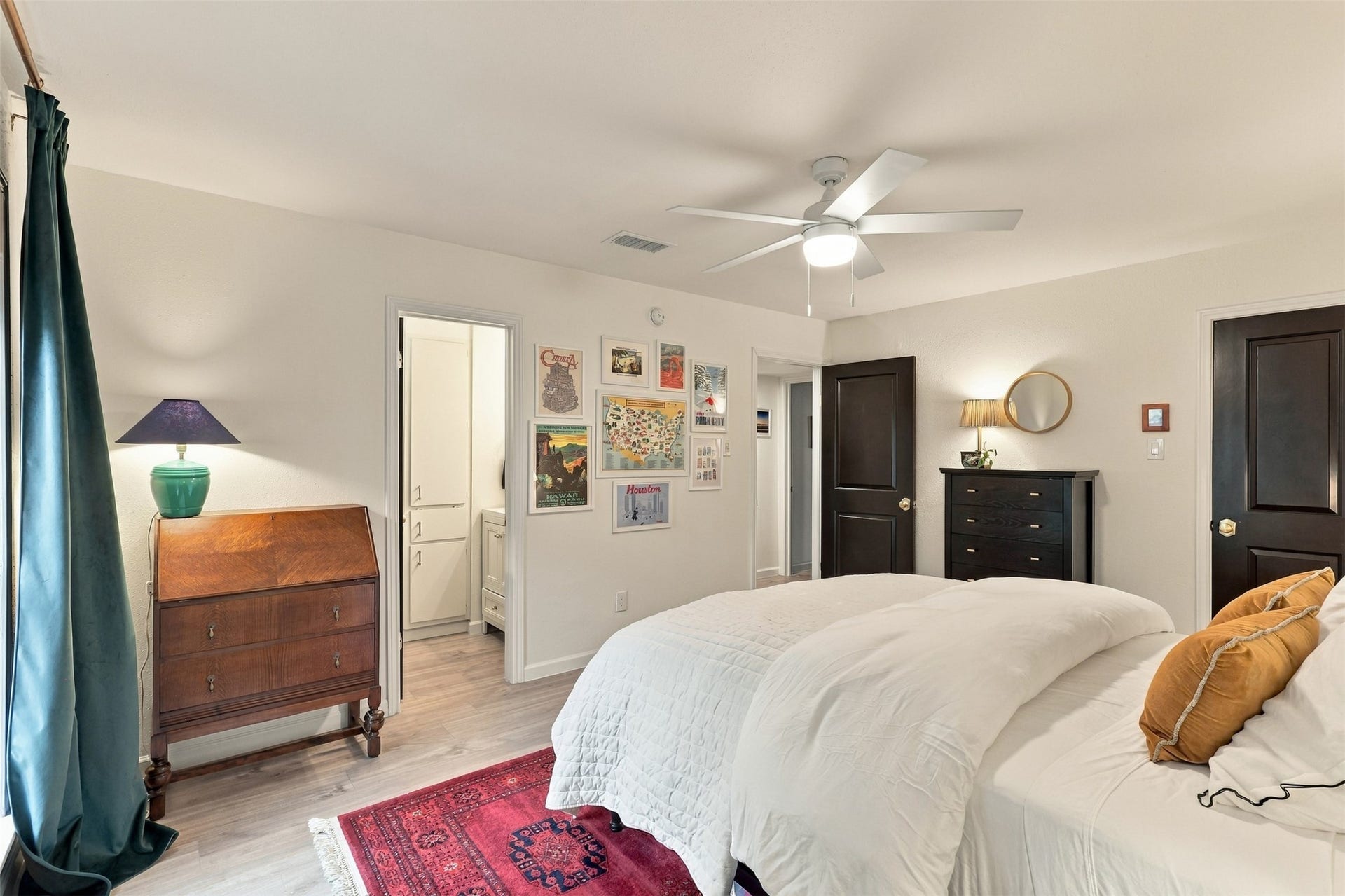 1201 Mcduffie Street, #212, Houston, TX 77019 Cozy bedroom featuring a stylish ceiling fan, elegant dark wood furniture, and a vibrant red area rug. Soft lighting complements the tasteful wall art, creating a warm and inviting atmosphere.