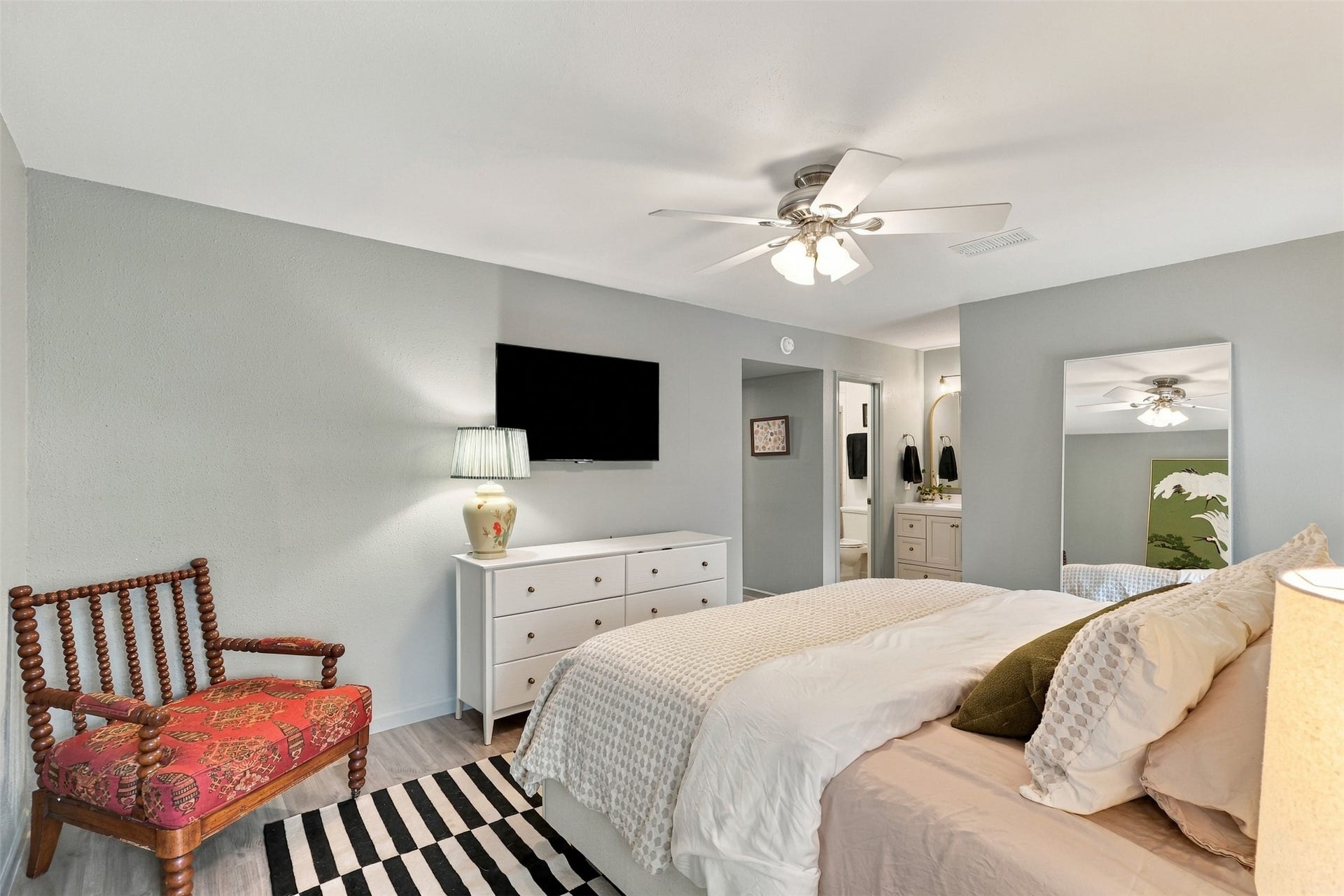 1201 Mcduffie Street, #212, Houston, TX 77019 Spacious bedroom with soft gray walls, featuring a cozy bed, a stylish chair, and a sleek dresser topped with a TV. A ceiling fan and ample lighting create a bright, welcoming atmosphere.