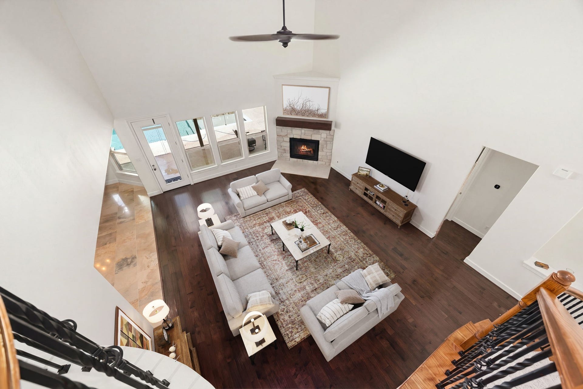 An aerial perspective from the upper landing highlights the dramatic volume of the two-story family room. This bird's-eye view beautifully captures the scale of the soaring ceilings, the placement of the corner stone fireplace, and the seamless transition between the rich hardwood floors and adjacent tiled flooring. *Virtually Staged.