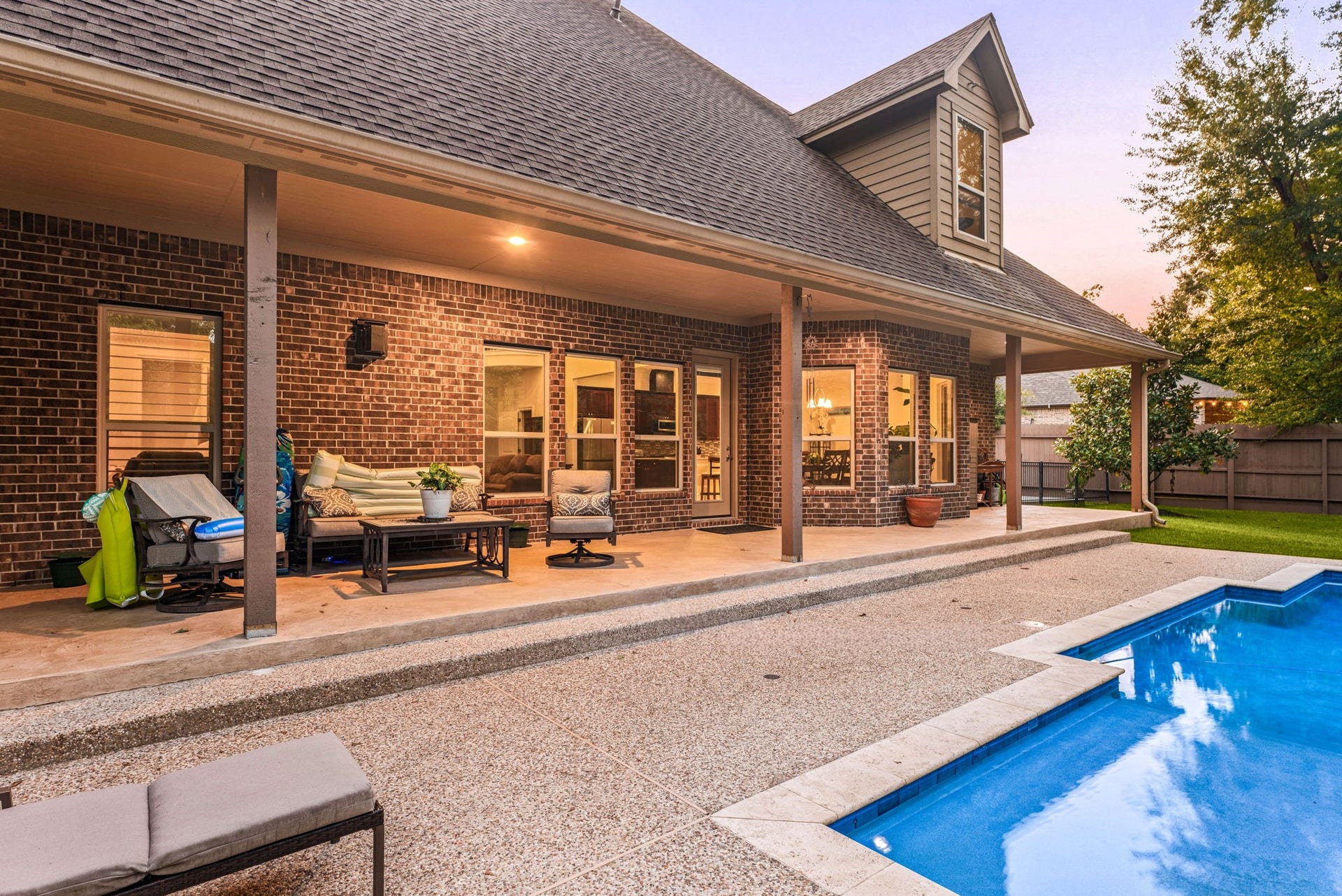 This covered back porch looks out over a beautiful in-ground swimming pool and hot tub. The space features a wooden ceiling with recessed lights, making it a perfect spot to relax in the shade while enjoying the private, tree-lined backyard.