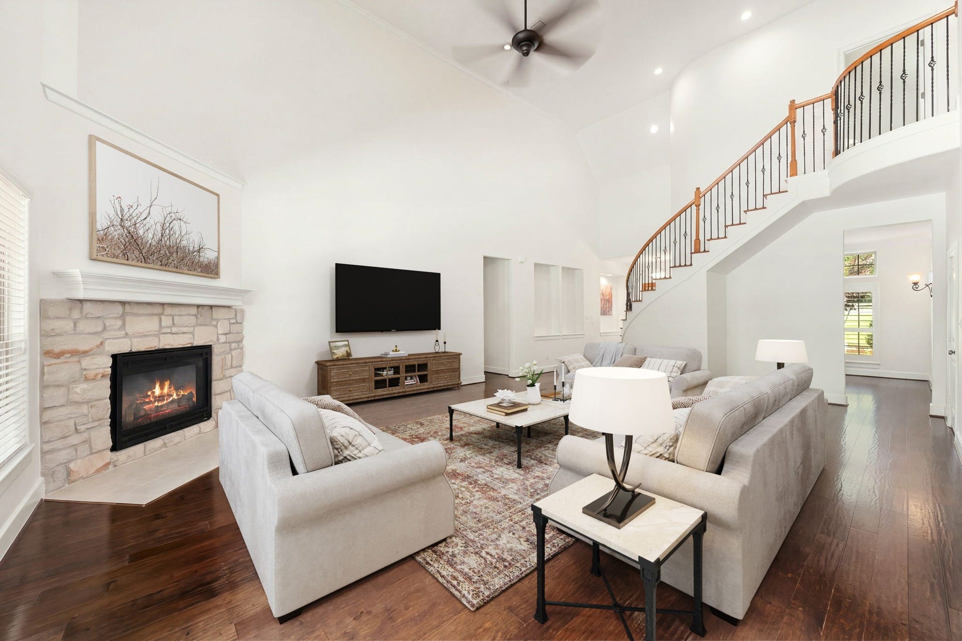 This alternative view of the grand family room highlights a curved staircase with elegant wrought-iron balusters as a striking architectural feature. The soaring vaulted ceilings, rich hardwood floors, and stone fireplace seamlessly connect to create a dramatic yet inviting living space. *Virtually Staged