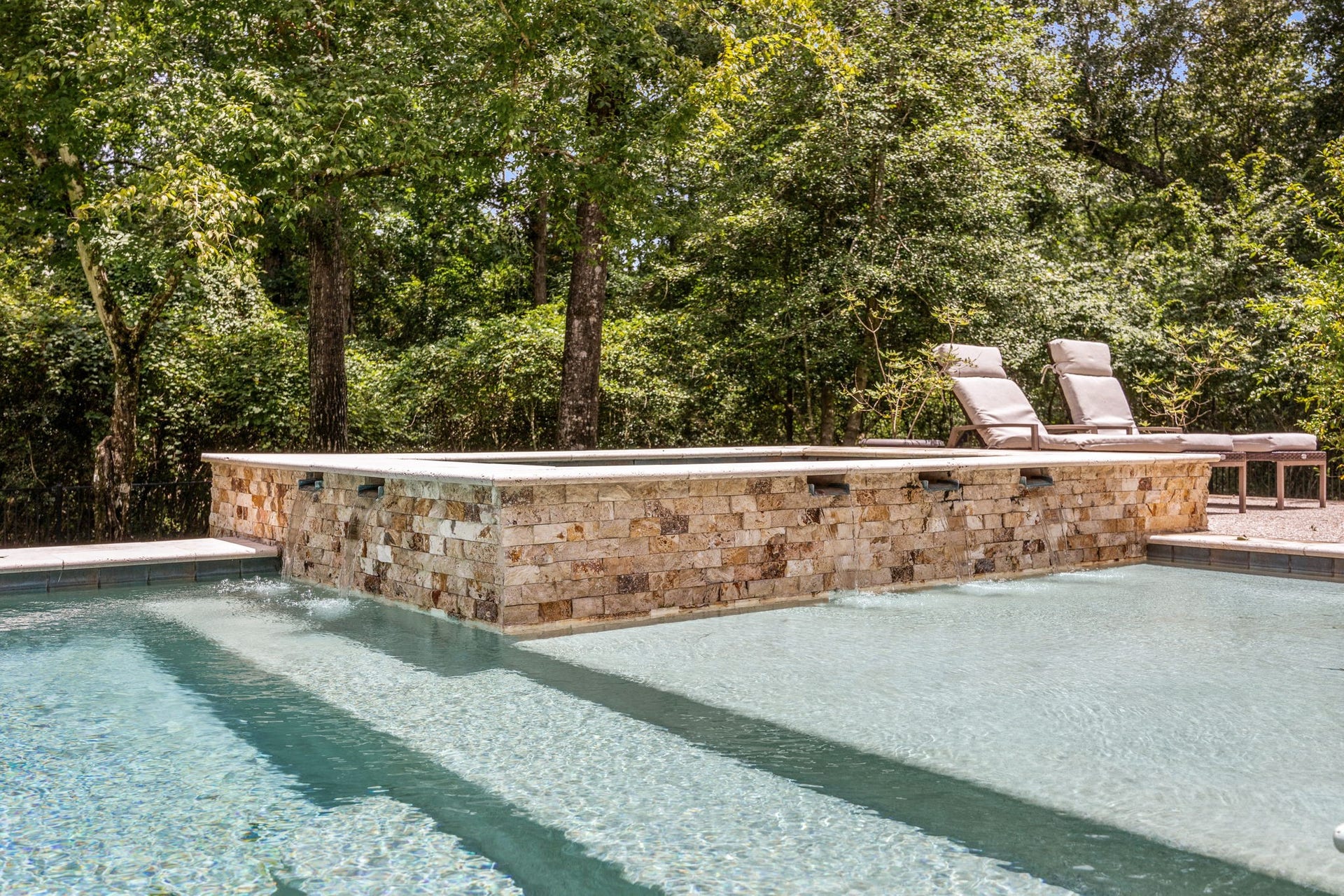 This crystal-clear in-ground swimming pool features a raised, stone-faced hot tub with multiple built-in water spillways that overflow gently into the pool below. The serene backyard oasis is surrounded by a dense wall of mature trees, offering absolute privacy.
