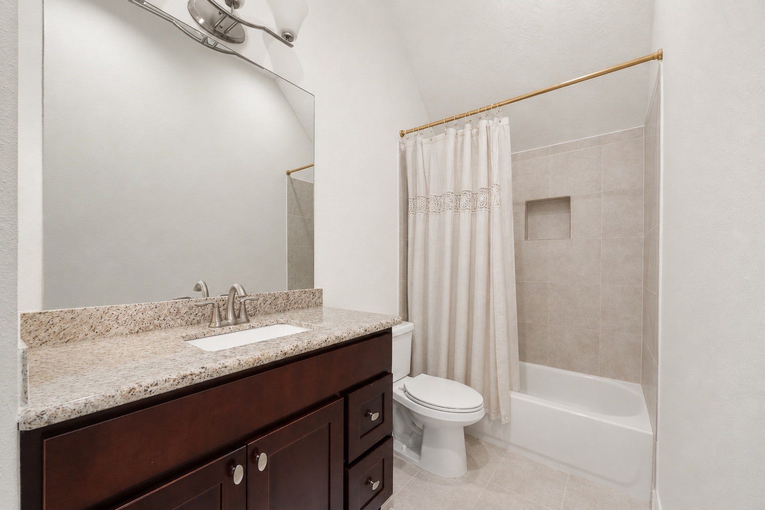 This pristine secondary bathroom offers a functional layout featuring a rich dark wood vanity with a premium granite countertop and an undermount sink. The space is complete with a clean tub-and-shower combination accented by a neutral tile surround and a convenient built-in shampoo niche.