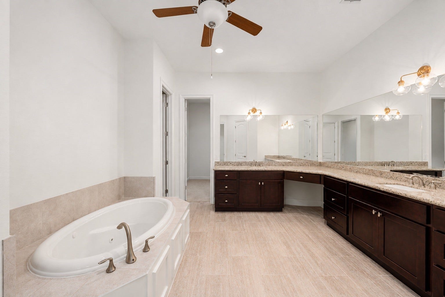 This spa-like primary bathroom features an expansive layout with an oversized soaking tub, wood-look tile flooring, and massive dual vanities. The rich dark cabinetry is topped with premium granite counters, offering endless storage and a dedicated sit-down makeup station beneath a wall of mirrors.