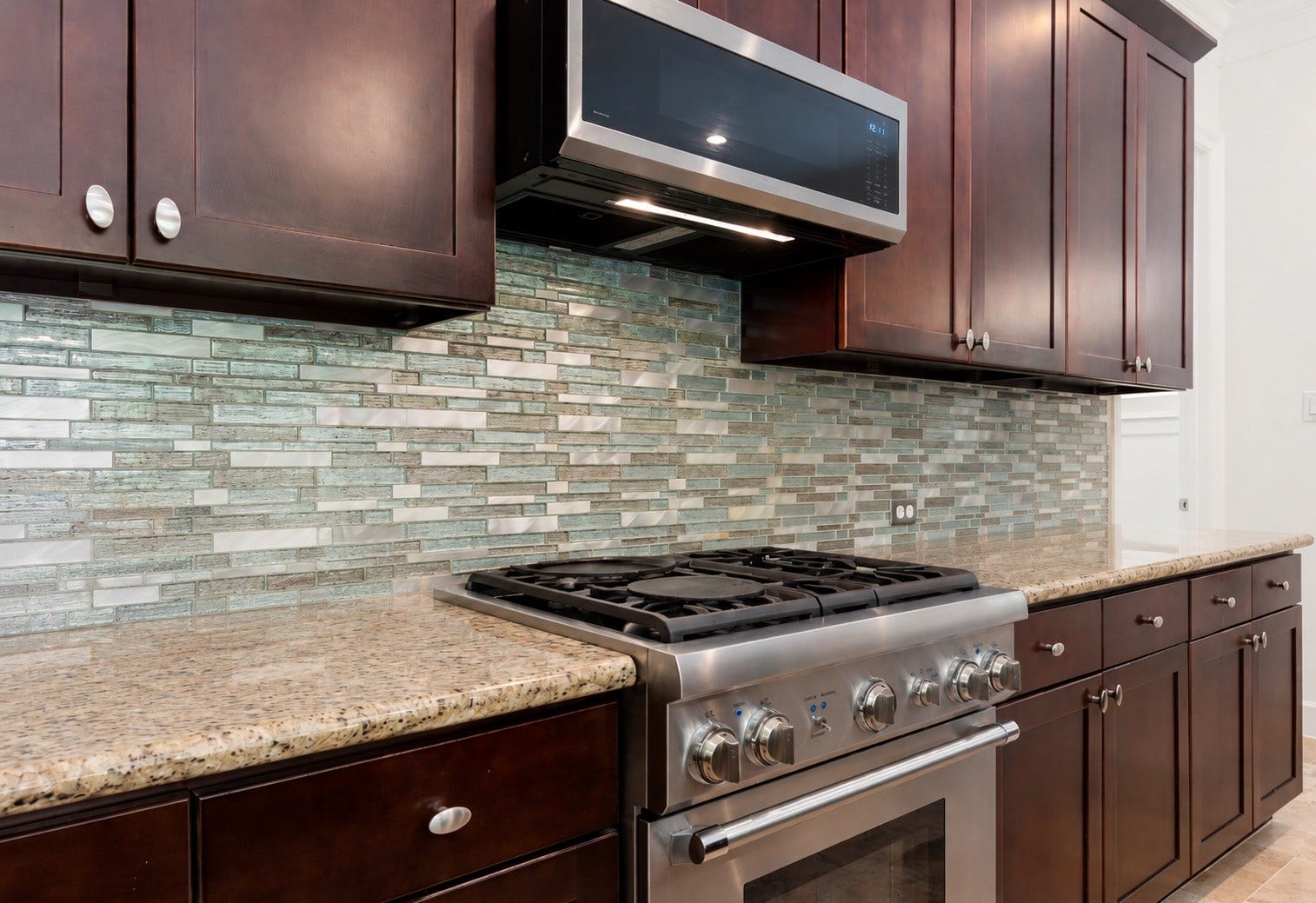 This close-up kitchen view highlights a professional-grade stainless steel gas range and a matching built-in microwave. The premium cooking station is beautifully framed by a stunning glass mosaic tile backsplash.