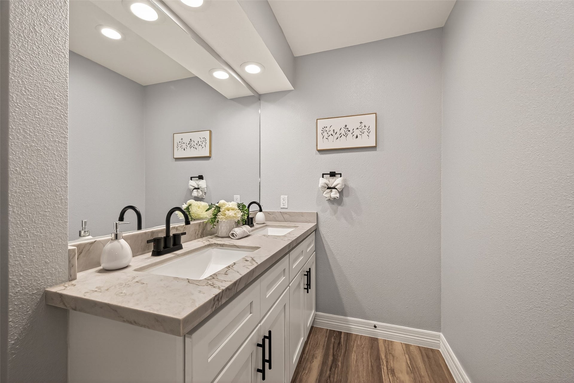 5422 Valerie Street, Bellaire, TX 77401 This bathroom features a modern design with a sleek vanity, marble countertop, and dual black faucets. Soft gray walls, wood-style flooring, and recessed lighting create a bright, inviting space.