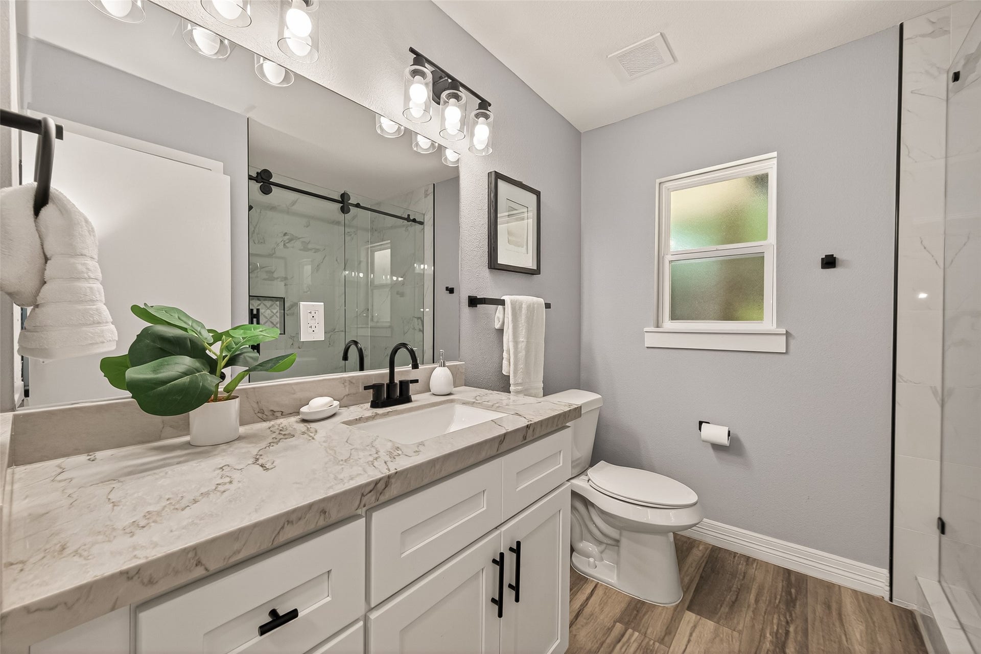 5422 Valerie Street, Bellaire, TX 77401 Modern bathroom with sleek vanity, marble countertop, and black fixtures. Features a glass-enclosed shower, wood-look flooring, and frosted window for privacy.