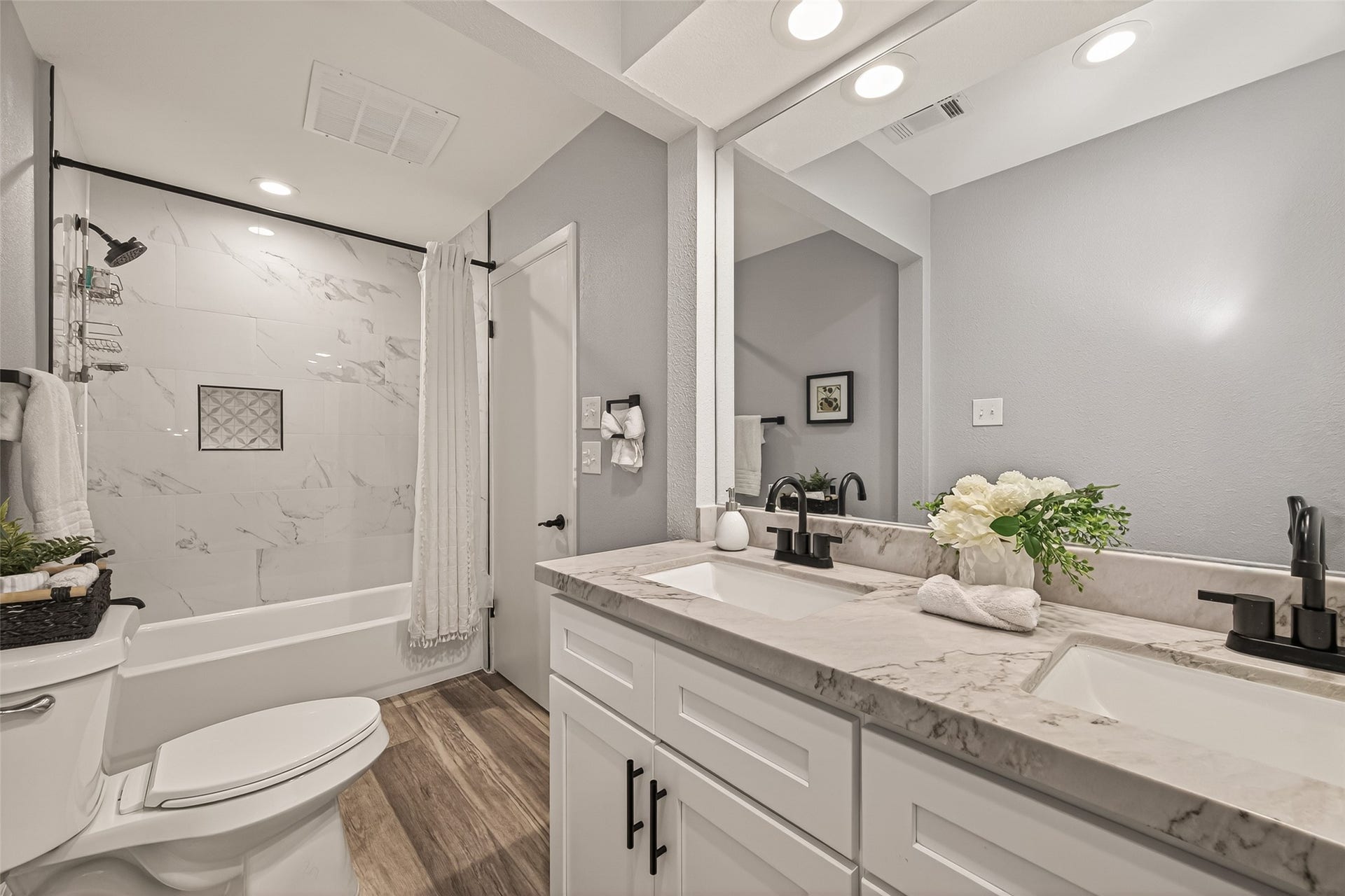 5422 Valerie Street, Bellaire, TX 77401 Secondary bathroom featuring a dual-sink vanity with marble countertop, sleek black fixtures, and abundant lighting. The tub-shower combination showcases elegant tile detailing, complemented by light wood-style flooring and neutral tones for a clean, inviting feel.