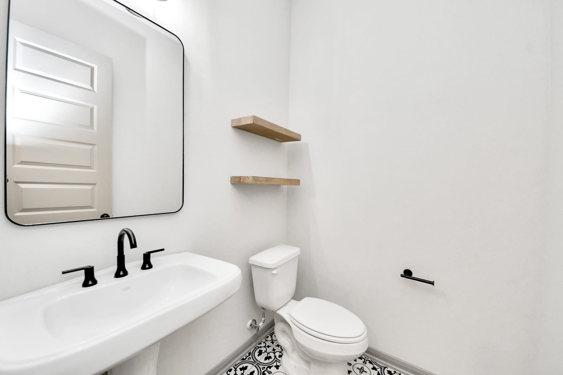 1423 Hackberry Heights Drive, Richmond, TX 77406 Chic half bathroom featuring a sleek pedestal sink with modern black fixtures, a stylish geometric-patterned floor, and minimalist wooden shelves.