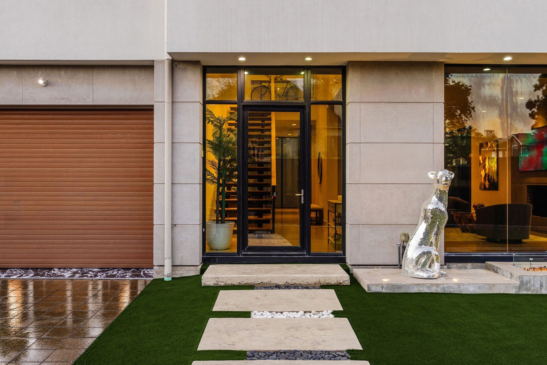 6002 Valley Forge Drive, Houston, TX 77057 Sleek modern entryway featuring a striking glass door, elegant stone pathway, and a contemporary sculpture, complemented by an adjacent garage with a warm-toned door.