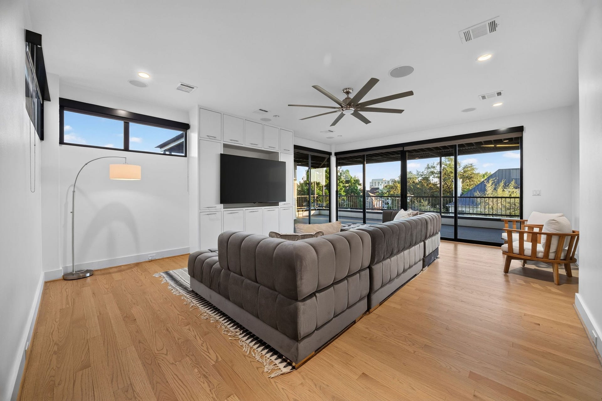 6002 Valley Forge Drive, Houston, TX 77057 Spacious living area with sleek hardwood flooring, a plush sectional sofa, and a large TV. Expansive windows and sliding doors offer abundant natural light and views of the greenery outside.