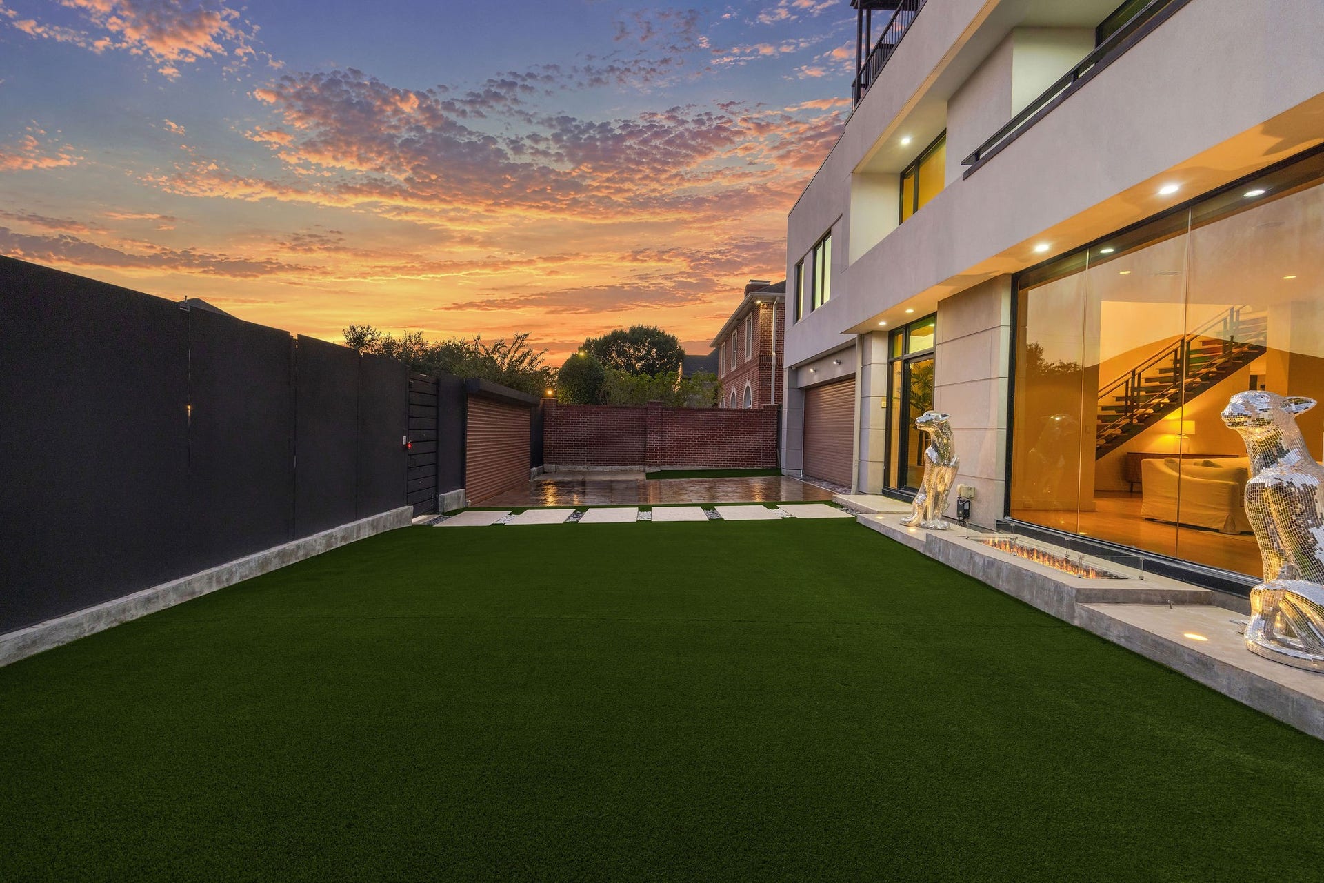 6002 Valley Forge Drive, Houston, TX 77057 Experience modern elegance with this stunning home's expansive patio, highlighted by sleek glass doors, lush artificial turf, and striking metallic sculptures, all set against a breathtaking sunset.